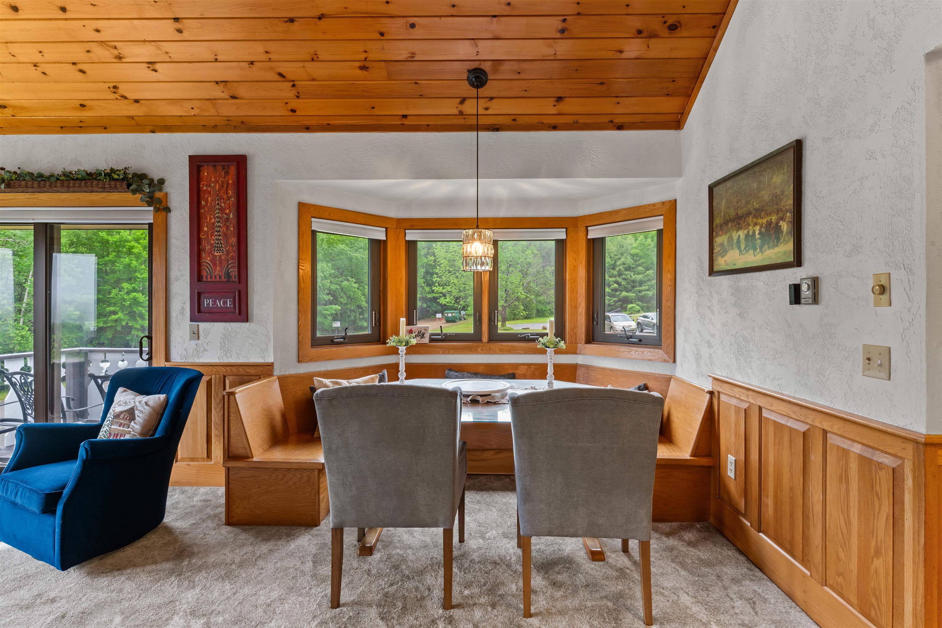 Dining nook with a built-in wooden bench, bay windows overlooking trees, and a wood-plank ceiling.