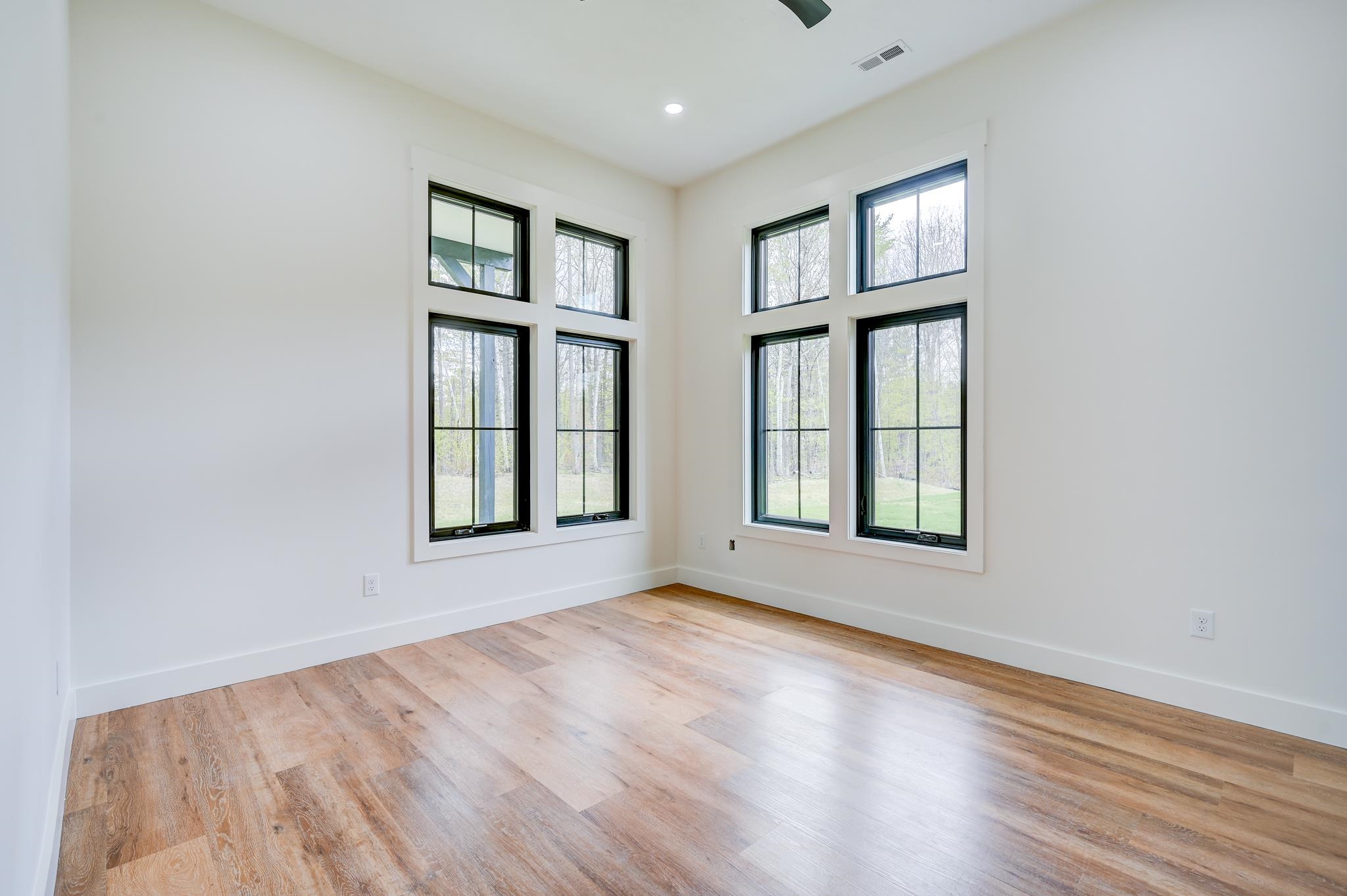 Empty room with wood-look flooring and tall windows overlooking a lawn and trees.