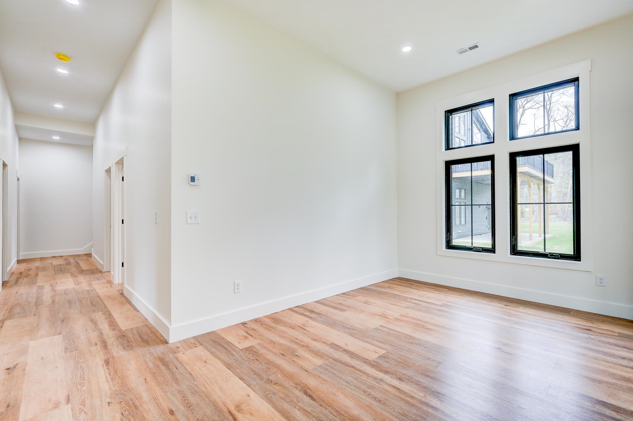 Vacant room with wood-look floors, tall black-framed windows, and a hallway opening to the left.