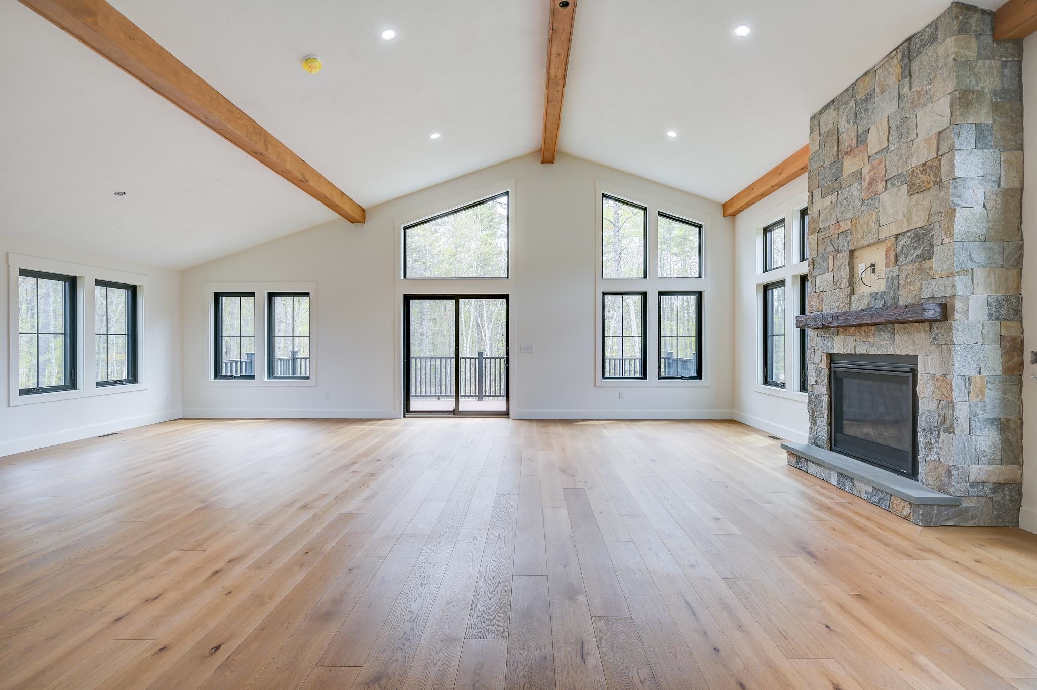 Vacant living room with a vaulted timber-beam ceiling, tall woodland-facing windows, and a stone fireplace.