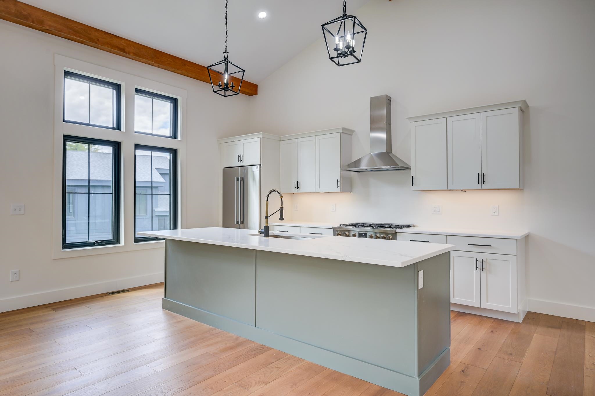 Modern kitchen with a large island, stainless steel appliances, tall windows, and exposed wood ceiling beam.
