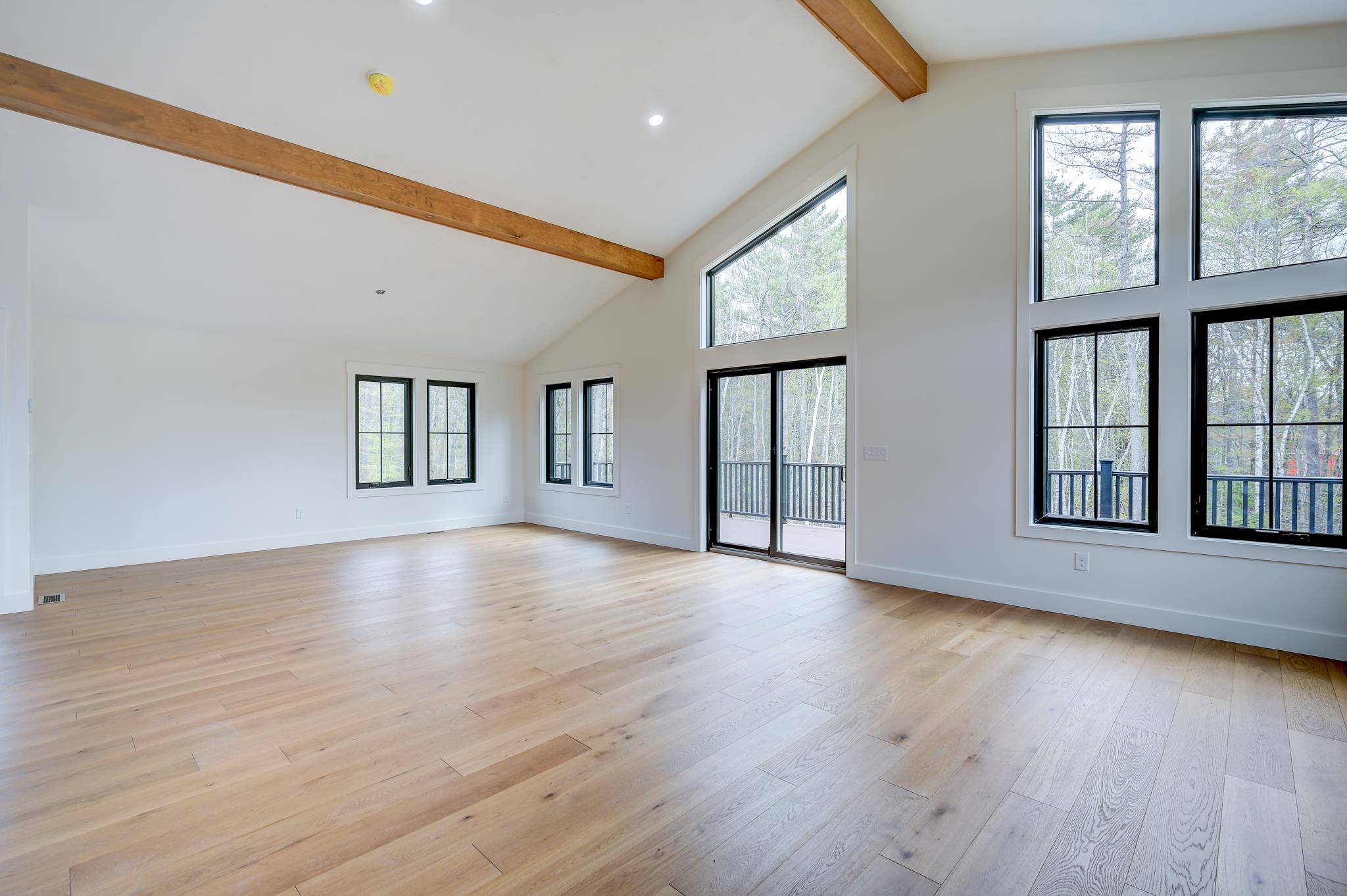 Vacant living room with vaulted ceiling, exposed wood beams, hardwood floors, and tall windows overlooking trees.