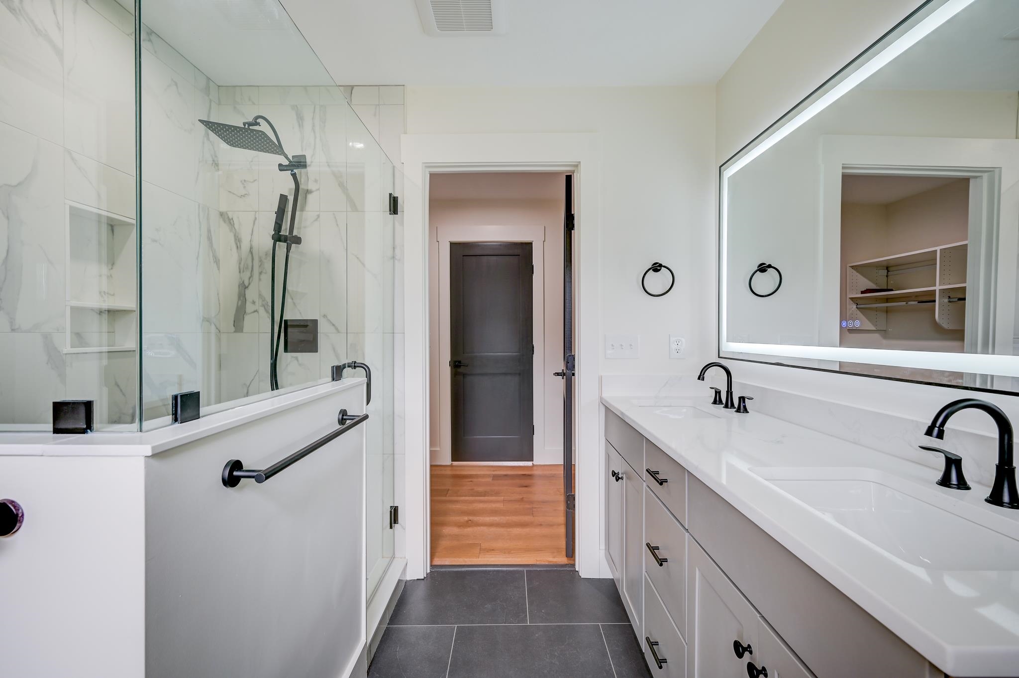 Modern bathroom with glass shower, marble-look tile, double vanity, and illuminated mirror.