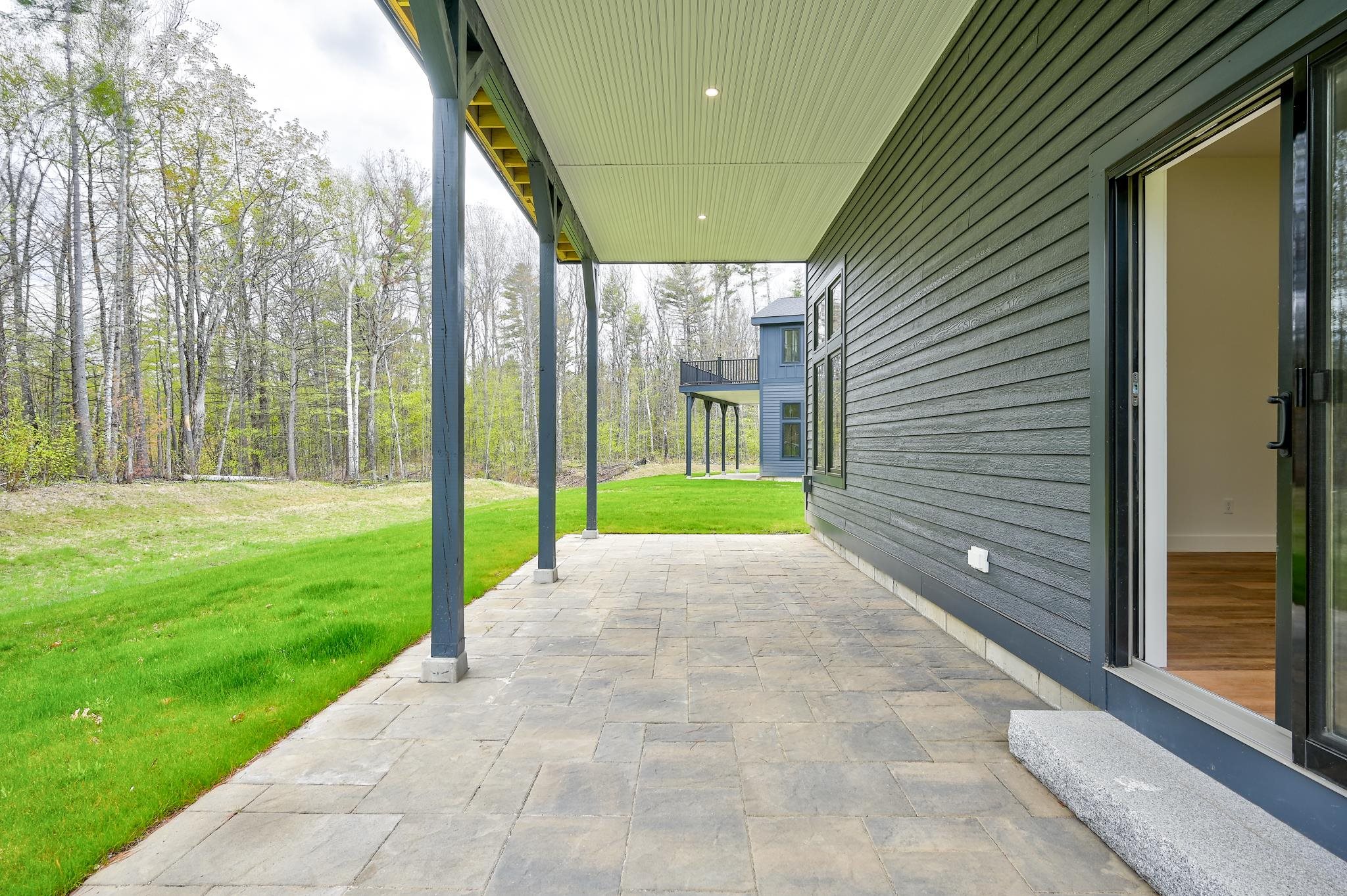 Covered stone patio beside a dark-sided home, overlooking a green lawn and woods.