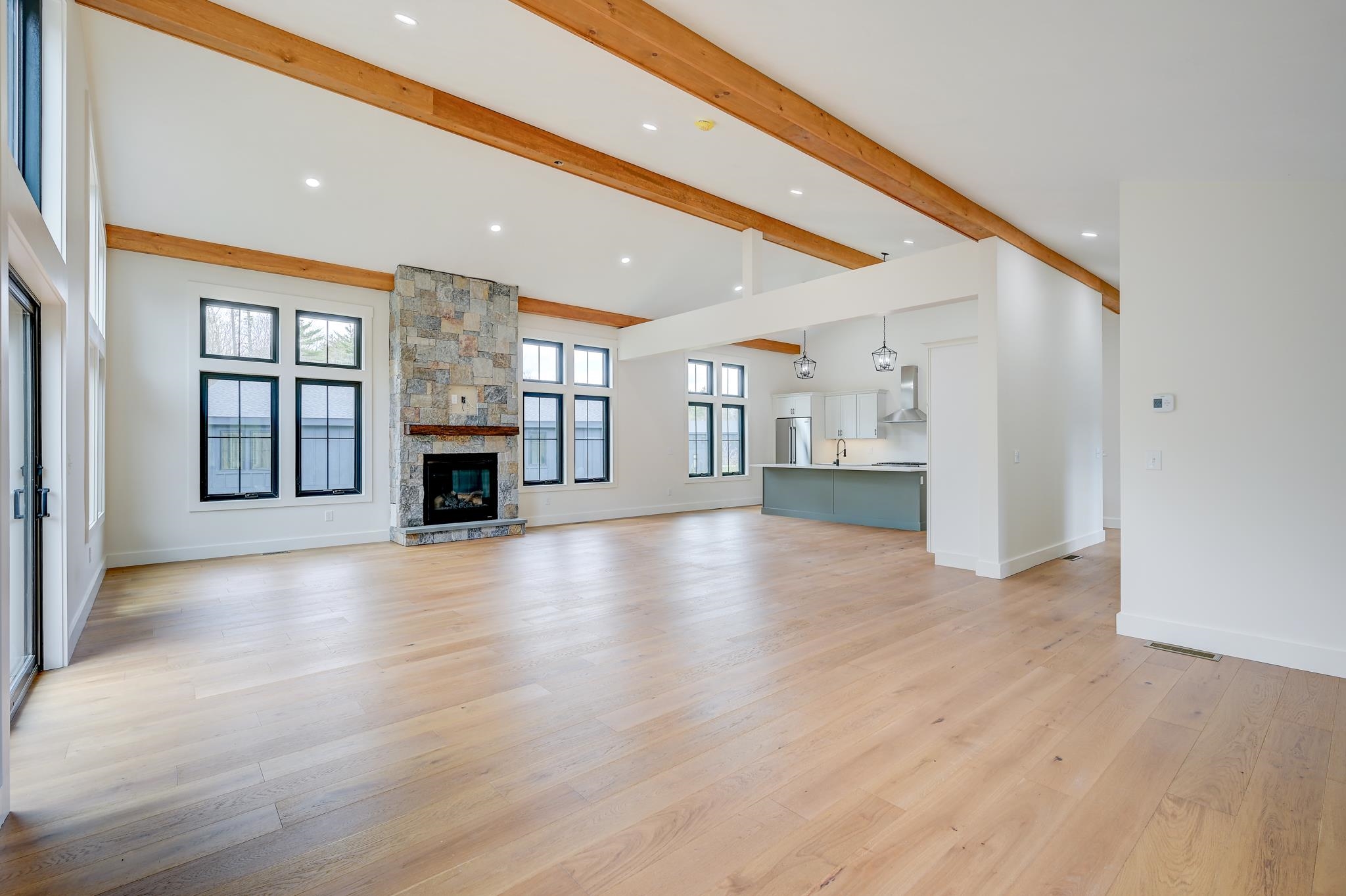 Open living room and kitchen with exposed timber beams, a stone fireplace, and pale hardwood floors.