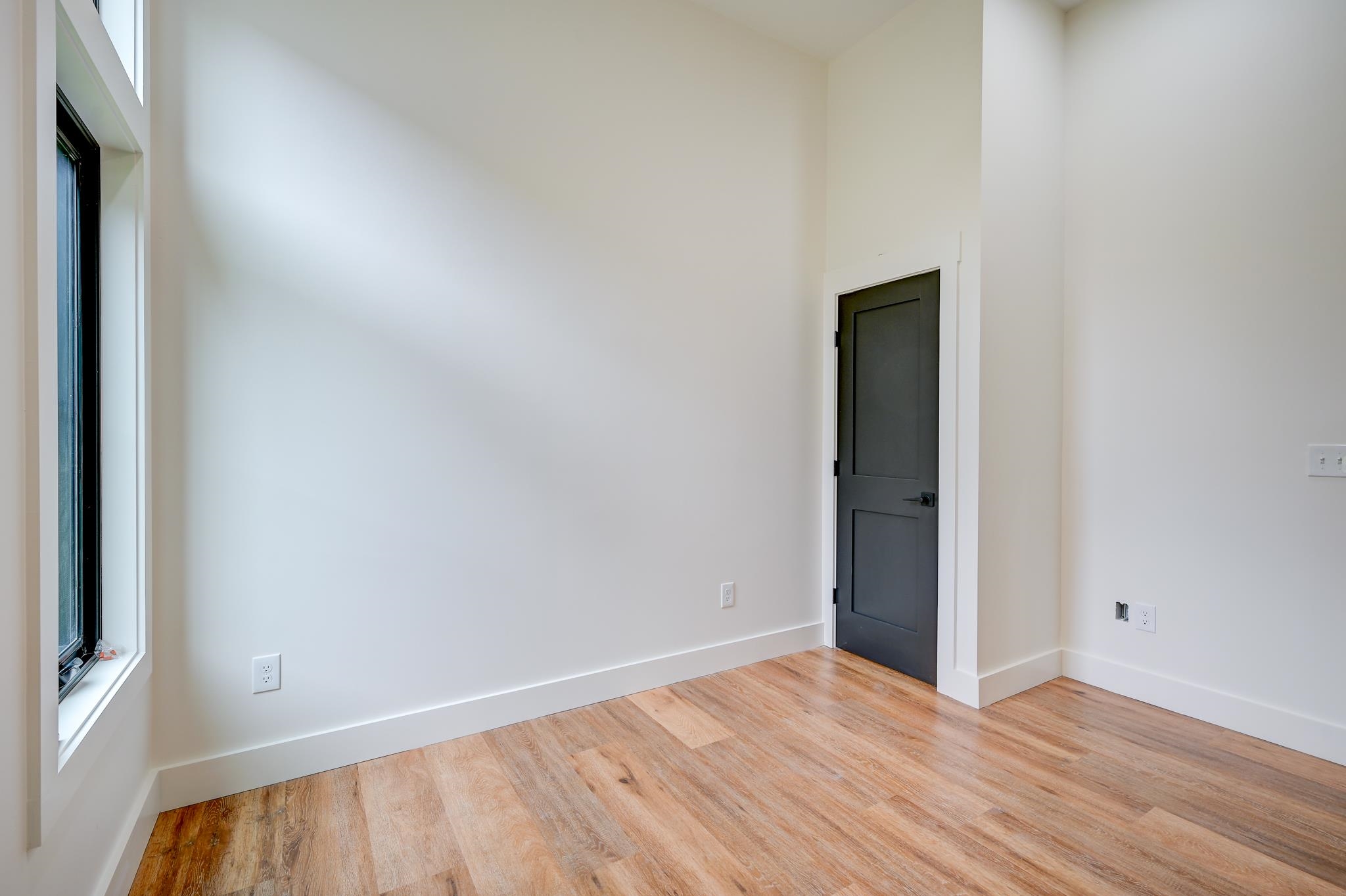 Empty room with a tall window, dark paneled door, and light wood flooring.