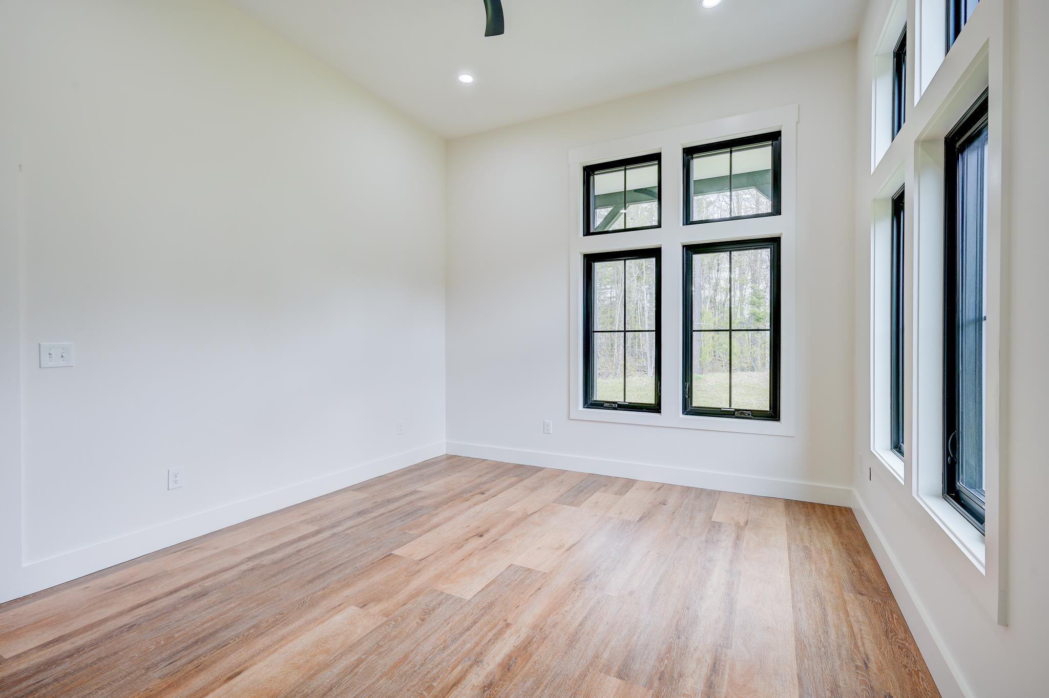 Empty room with light wood flooring and tall black-framed windows overlooking trees.