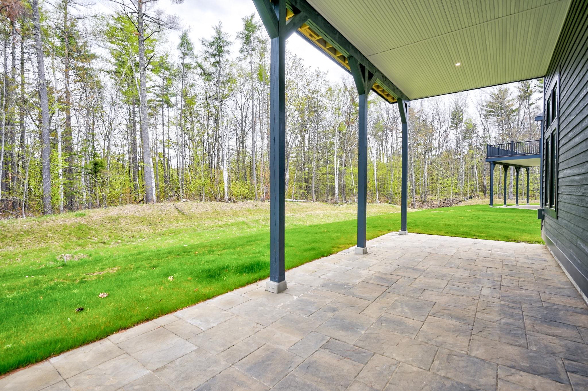 Covered stone patio beside a dark-sided home, overlooking a green lawn and wooded yard.