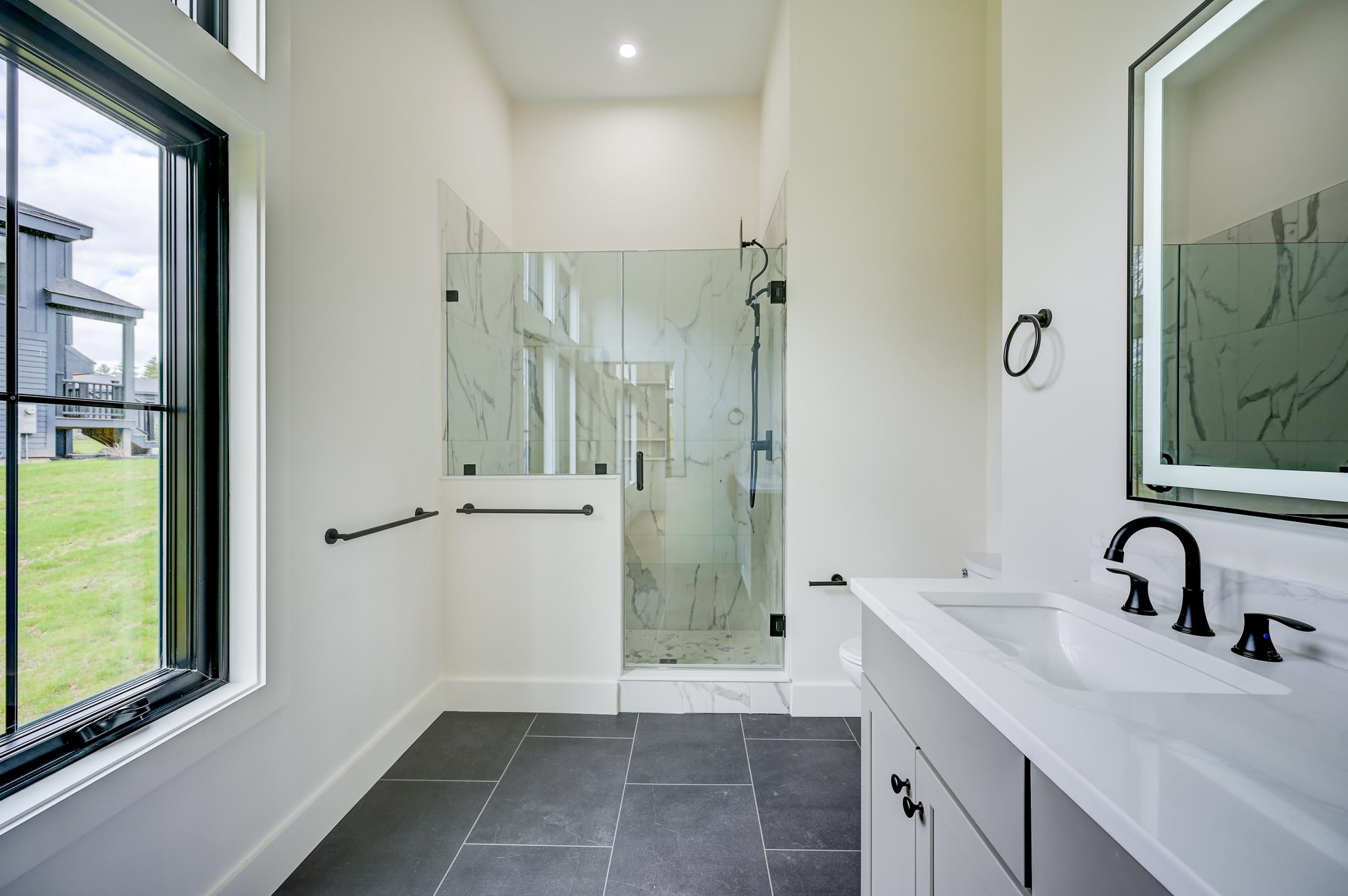 Bathroom with a marble-look glass shower, double-sink vanity, black faucets, and a large window.