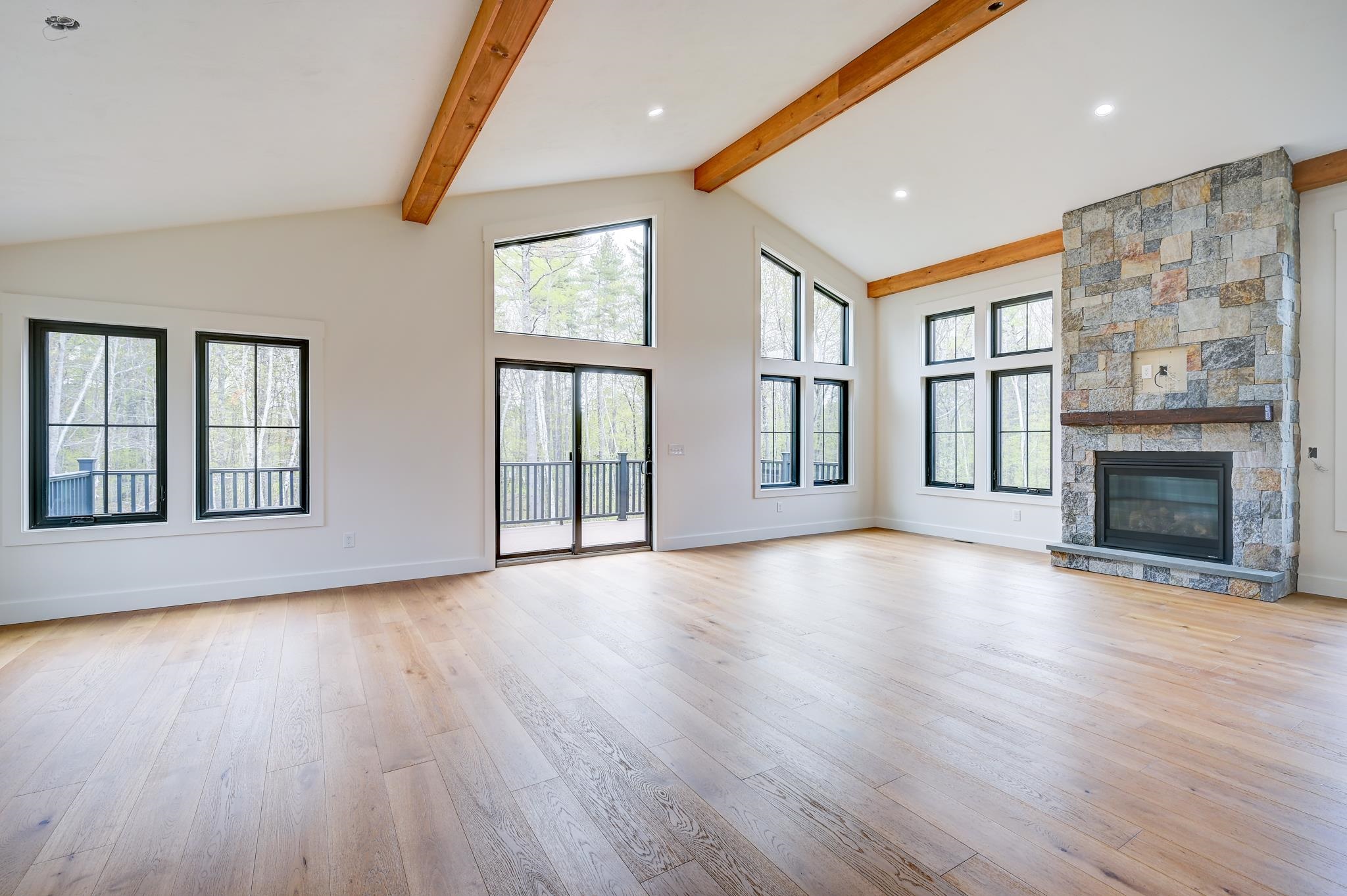 Vacant living room with vaulted timber-beam ceiling, stone fireplace, and large windows overlooking trees.