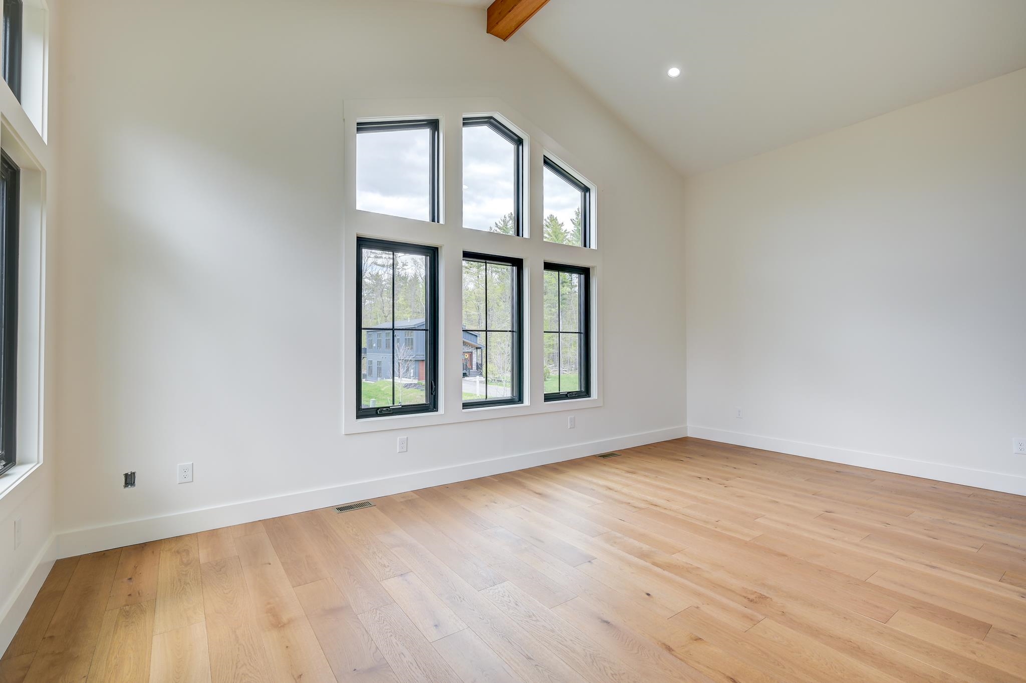 Empty room with vaulted ceiling, light hardwood floors, and tall windows overlooking trees.