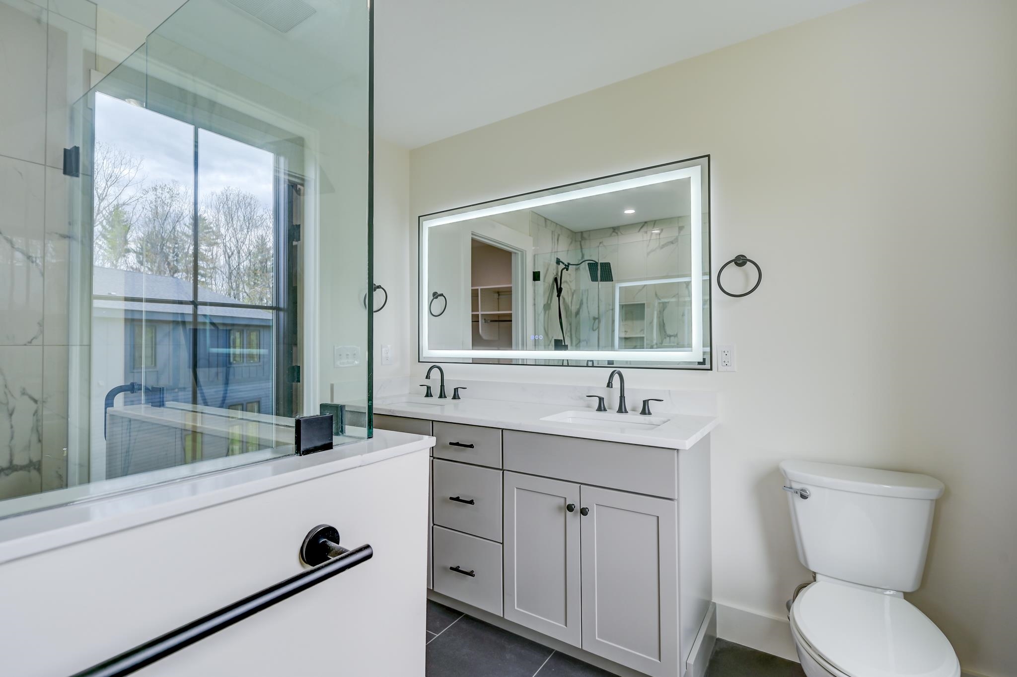 Modern bathroom with glass-enclosed tub, double-sink vanity, illuminated mirror, and black fixtures.