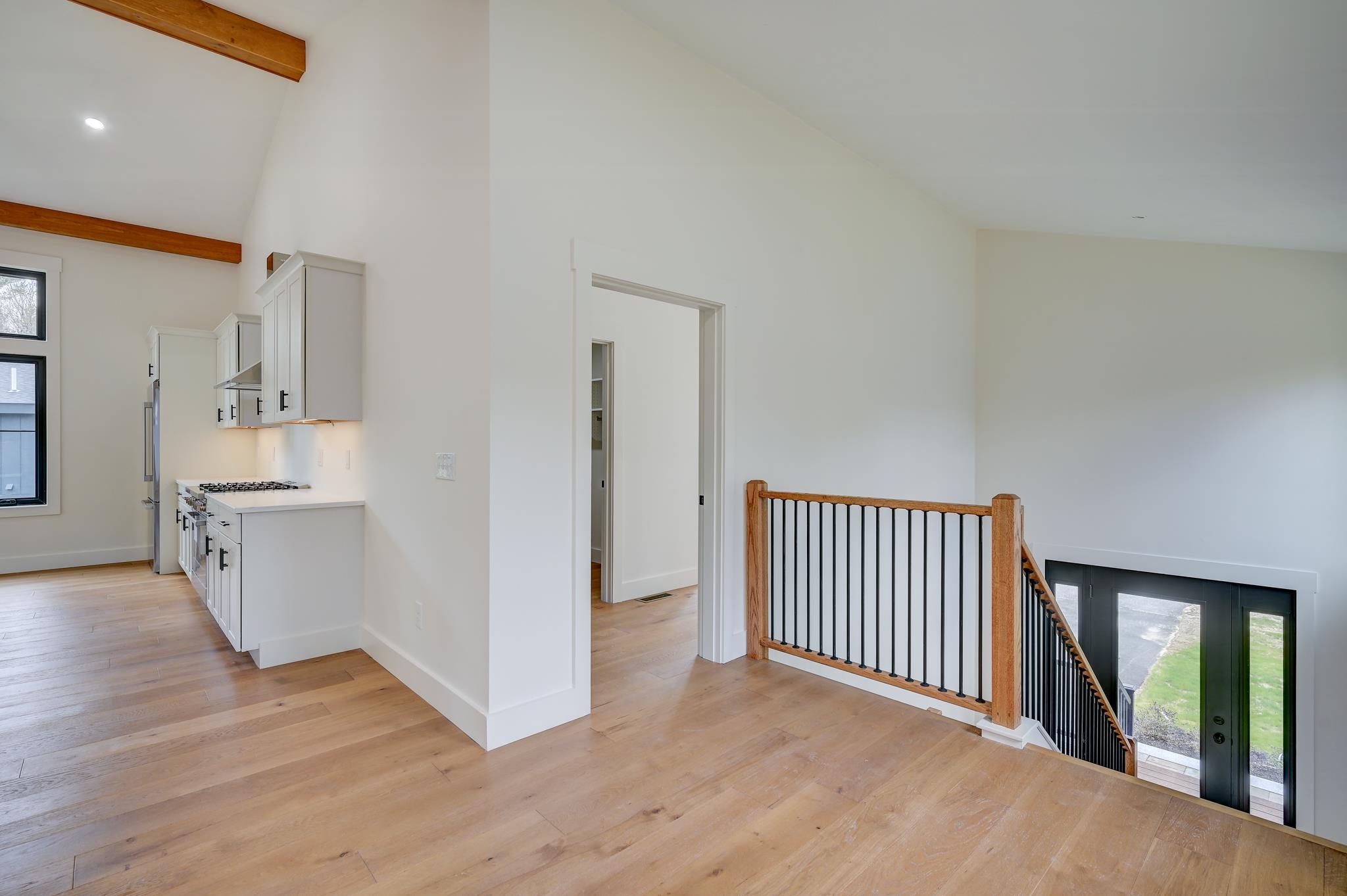 Open kitchen and stair landing with light wood floors, exposed beams, and a black-metal railing.