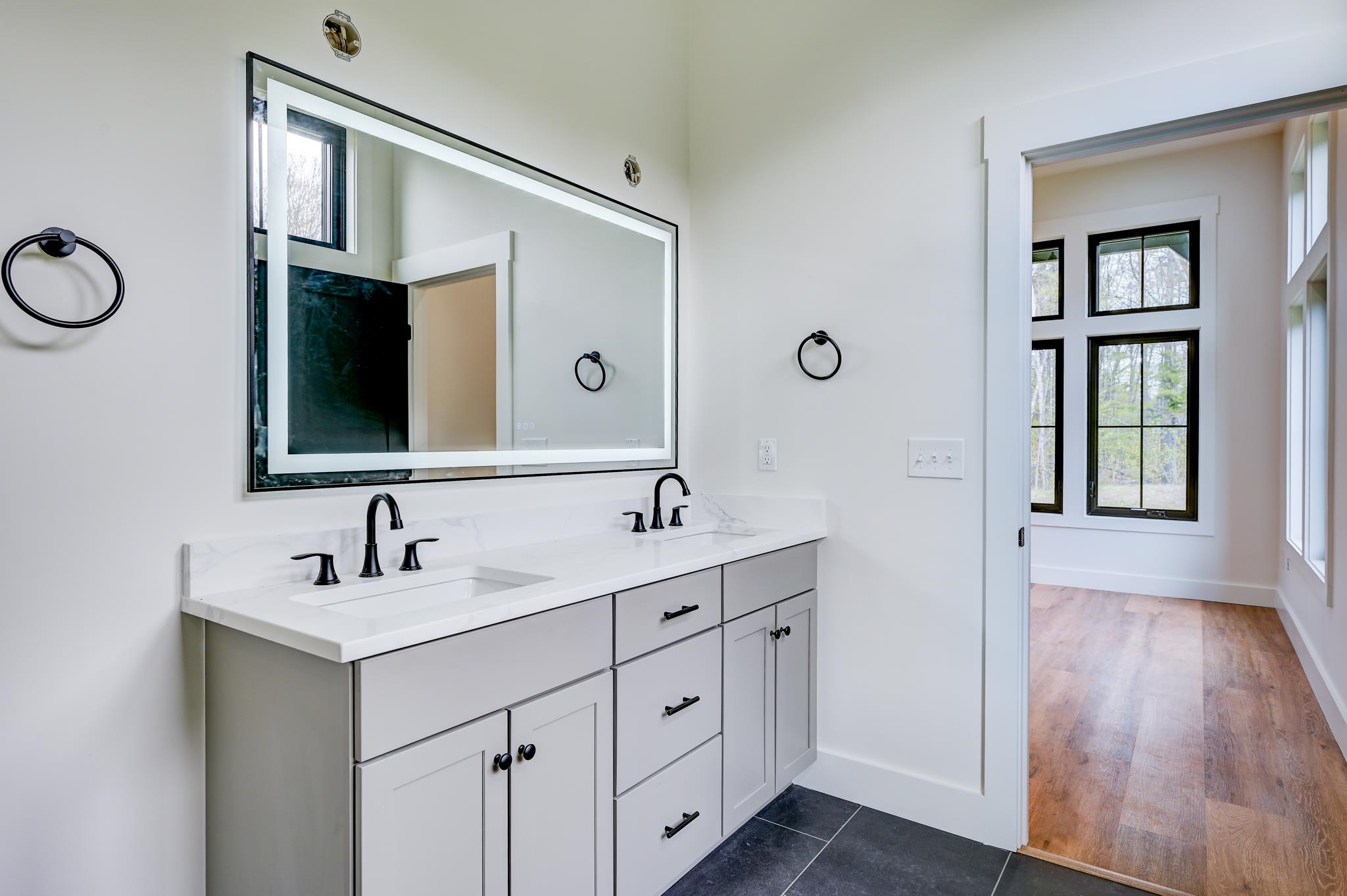 Bright bathroom with a white double vanity, black faucets, large mirror, and doorway to a sunlit room.