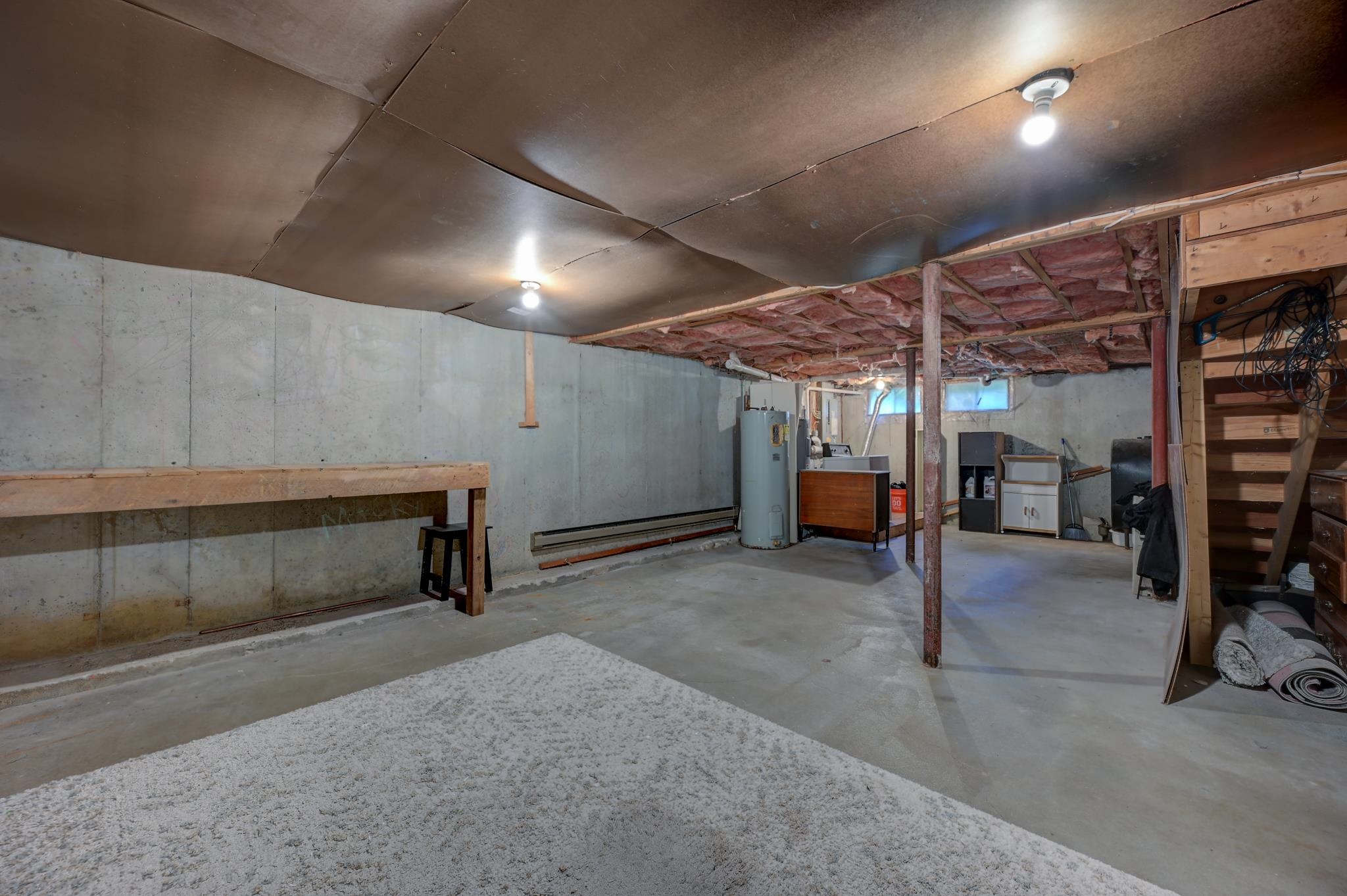 Basement with exposed ceiling insulation, a water heater, workbench, and storage shelving.