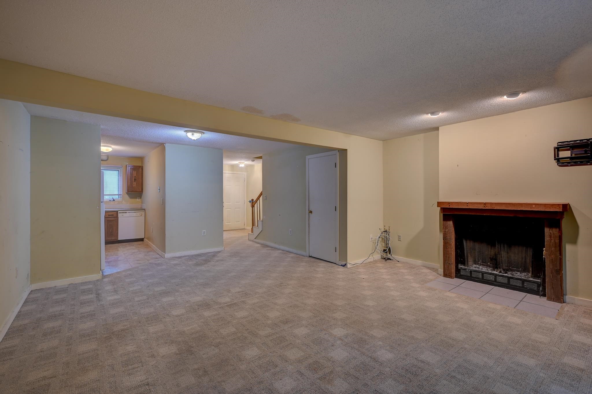 Unfurnished living room with beige carpet, a wood-framed fireplace, and a kitchen visible beyond.