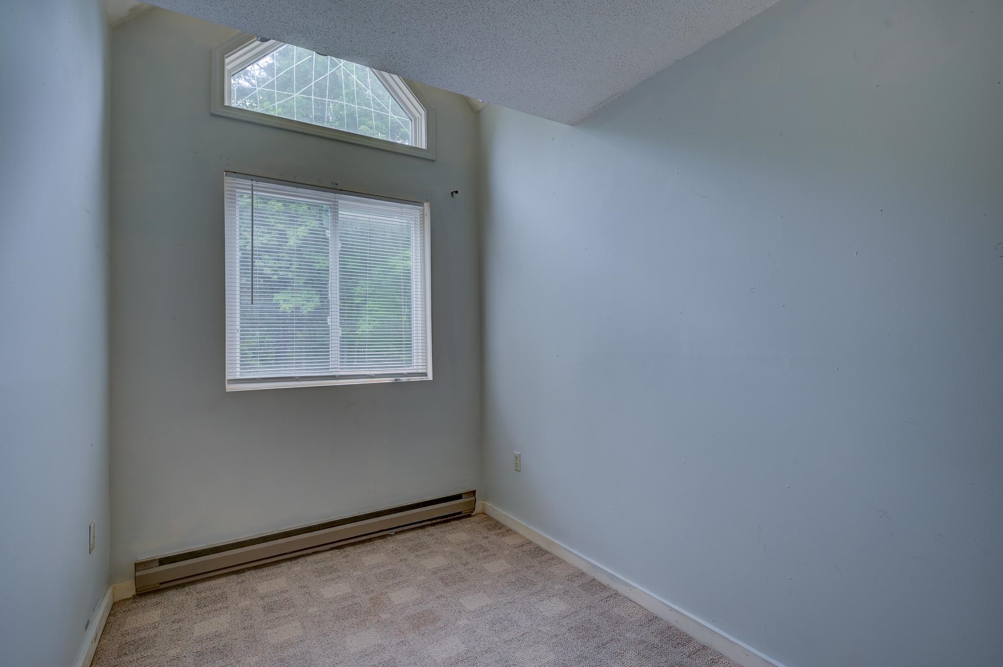 Empty carpeted room with a high angled ceiling, large window with blinds, and baseboard heater.
