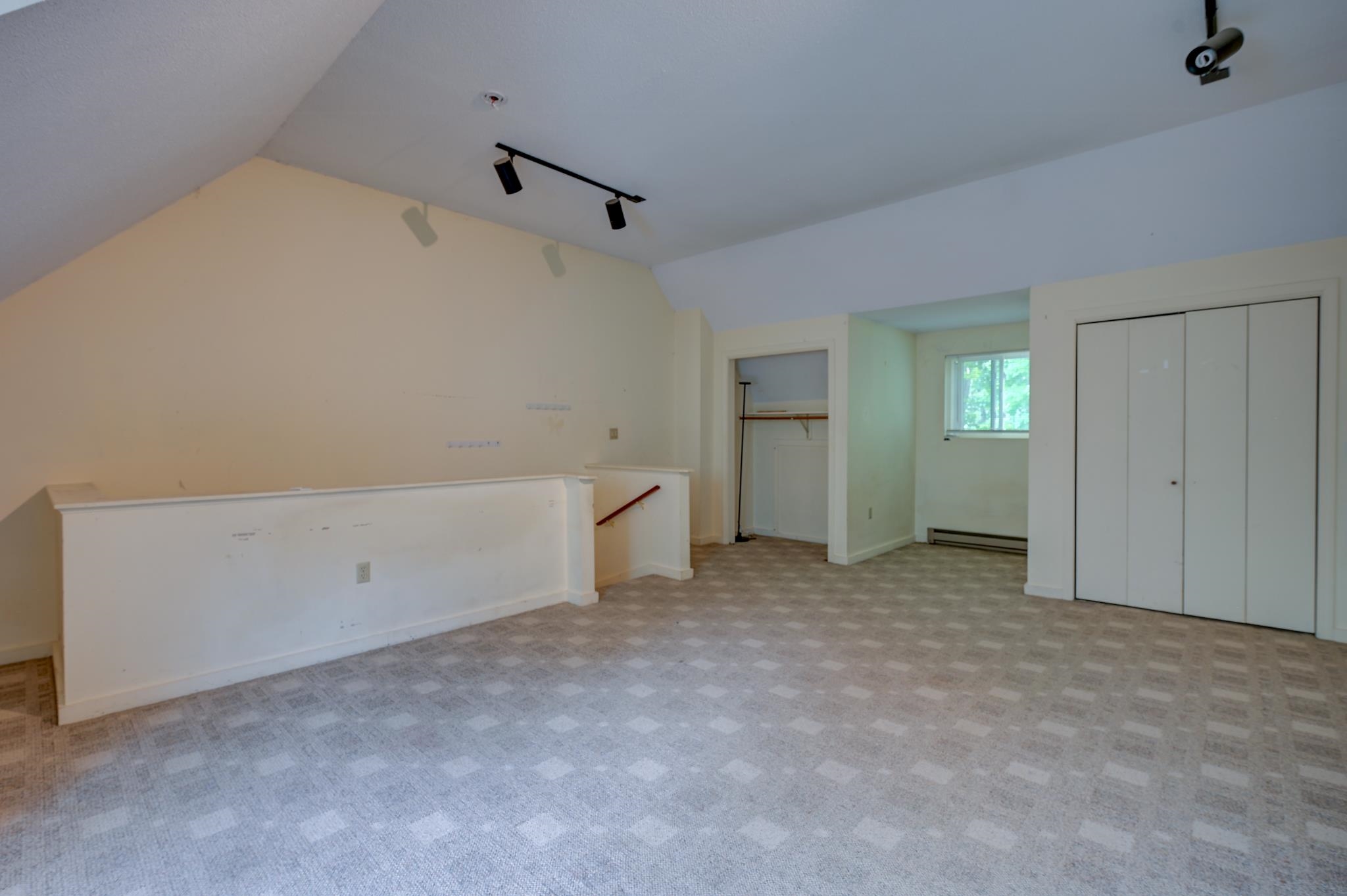 Empty carpeted room with a sloped ceiling, half wall, closet, and window overlooking greenery.