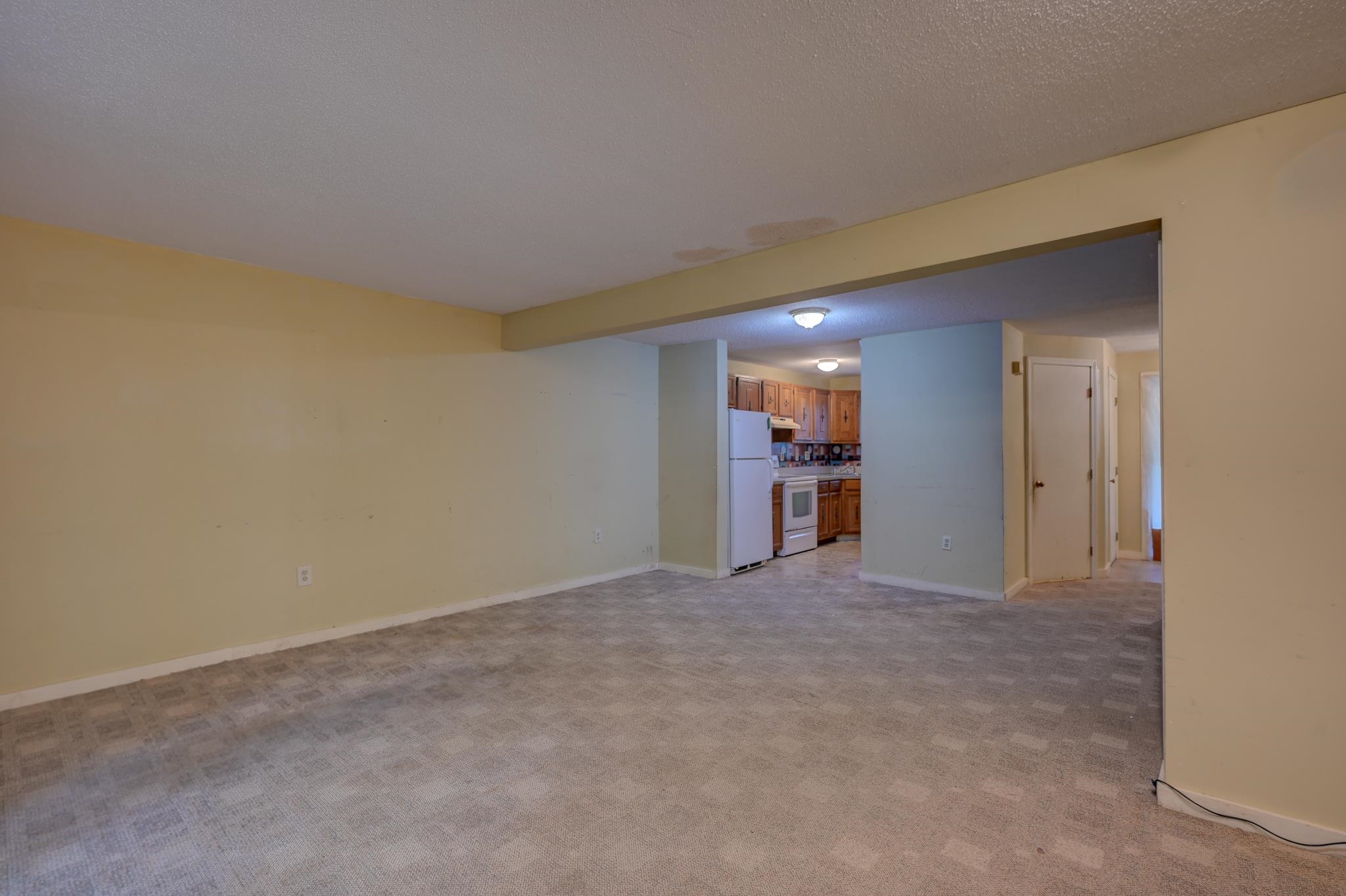 Empty living room with patterned carpet, open to a kitchen with wood cabinets and white appliances.