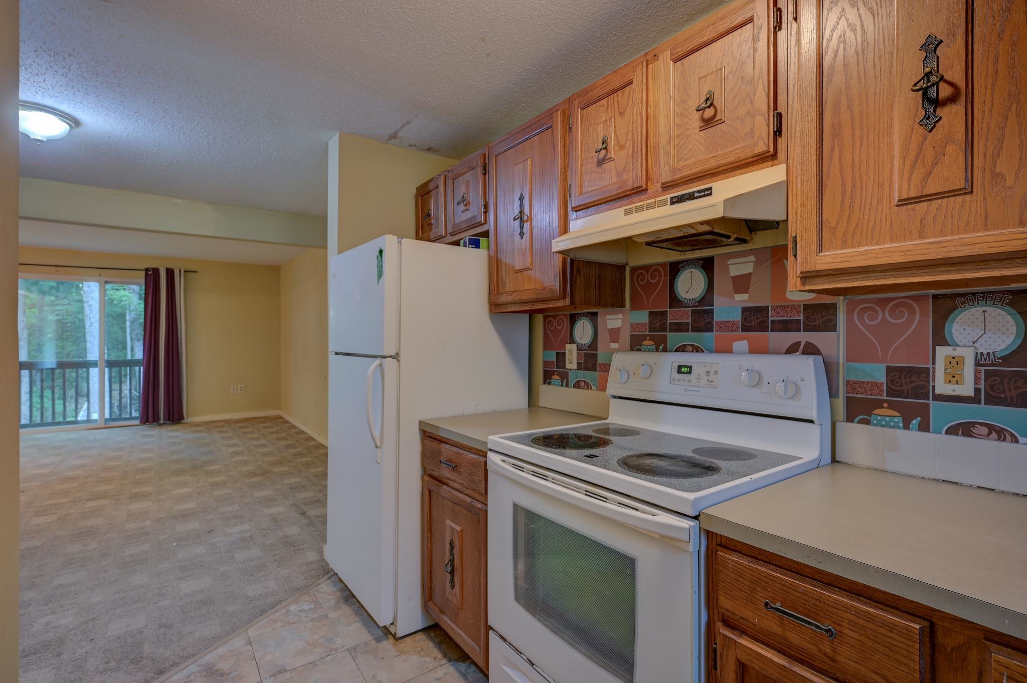 Kitchen with wood cabinets, white appliances, coffee-themed backsplash, and adjoining living area.