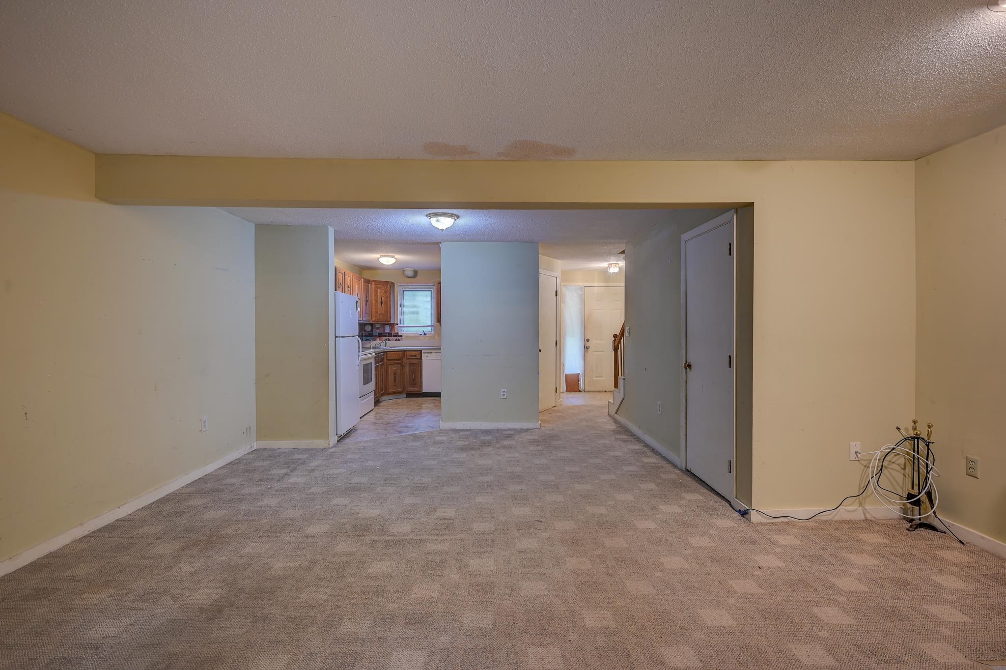 Empty carpeted living area looking toward the kitchen and entry hallway, with loose cables along the right wall.
