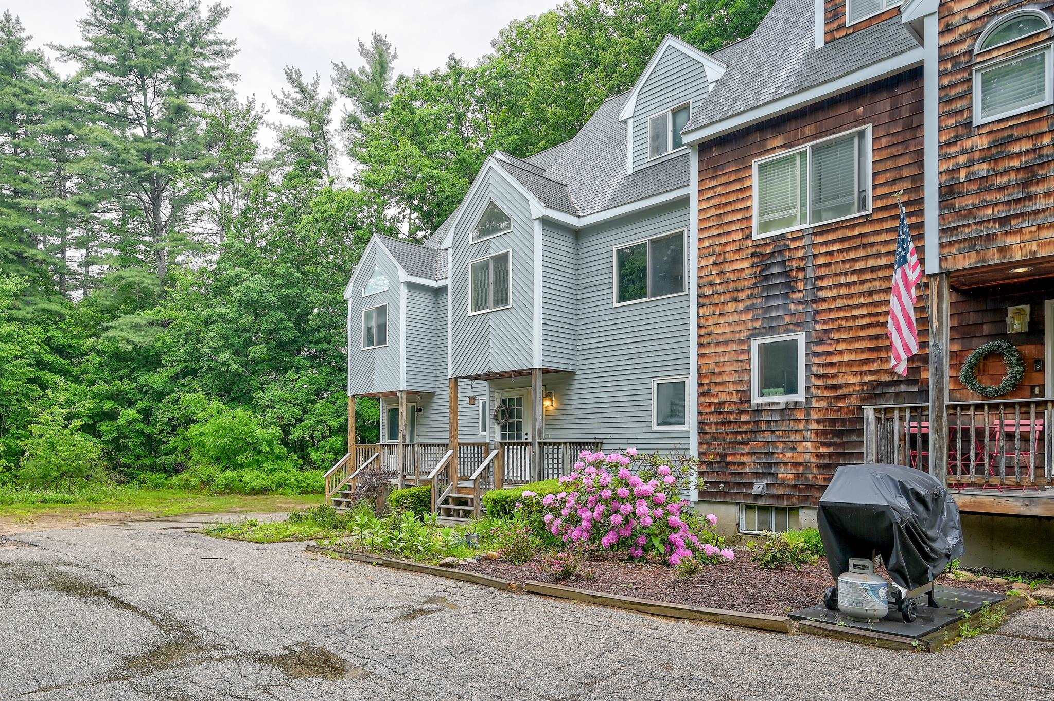 Residential building exterior with gray and brown siding, entry porches, and pink flowering shrubs.