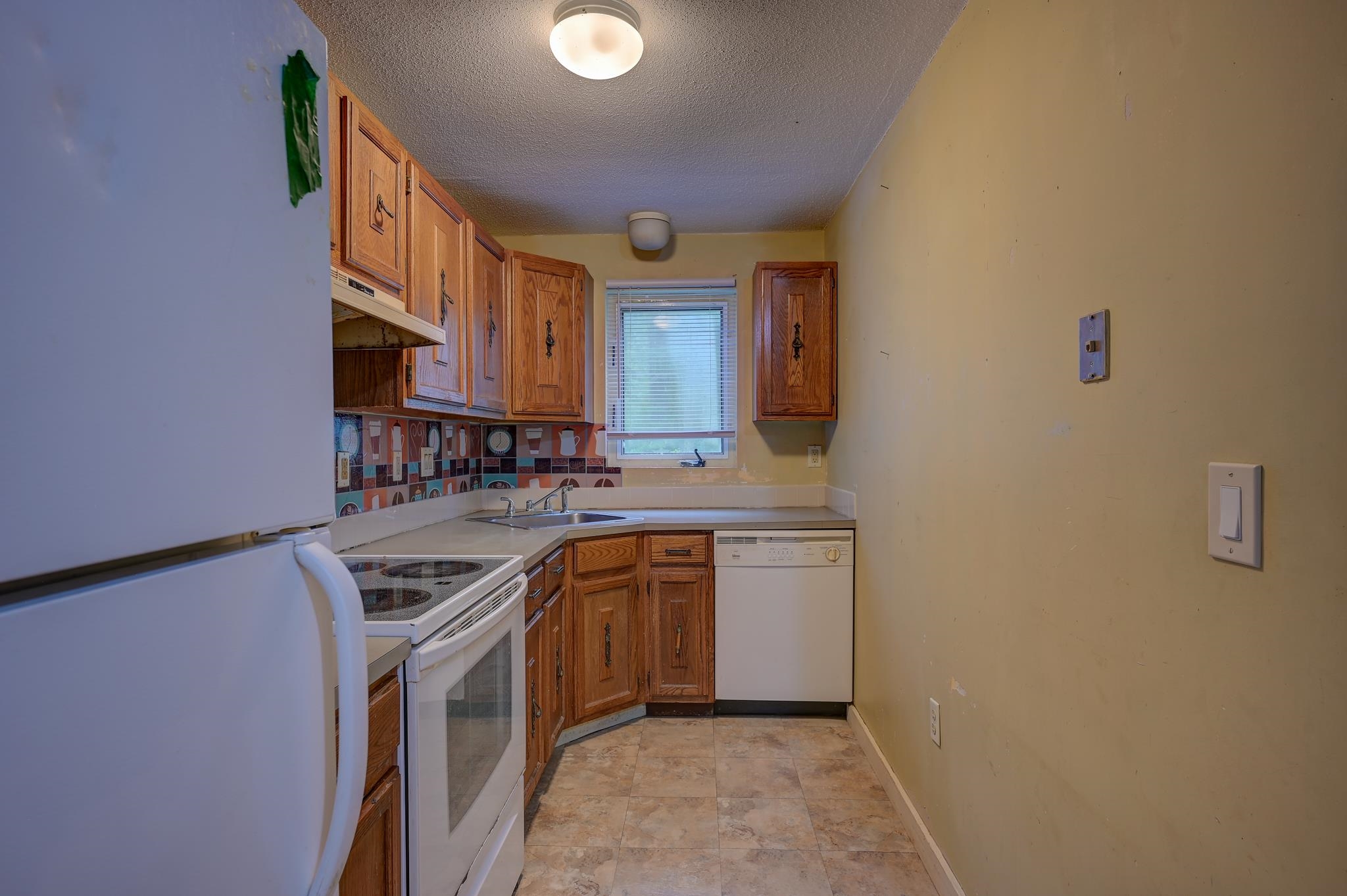 Narrow kitchen with wood cabinets, white appliances, patterned backsplash, and a window above the sink.
