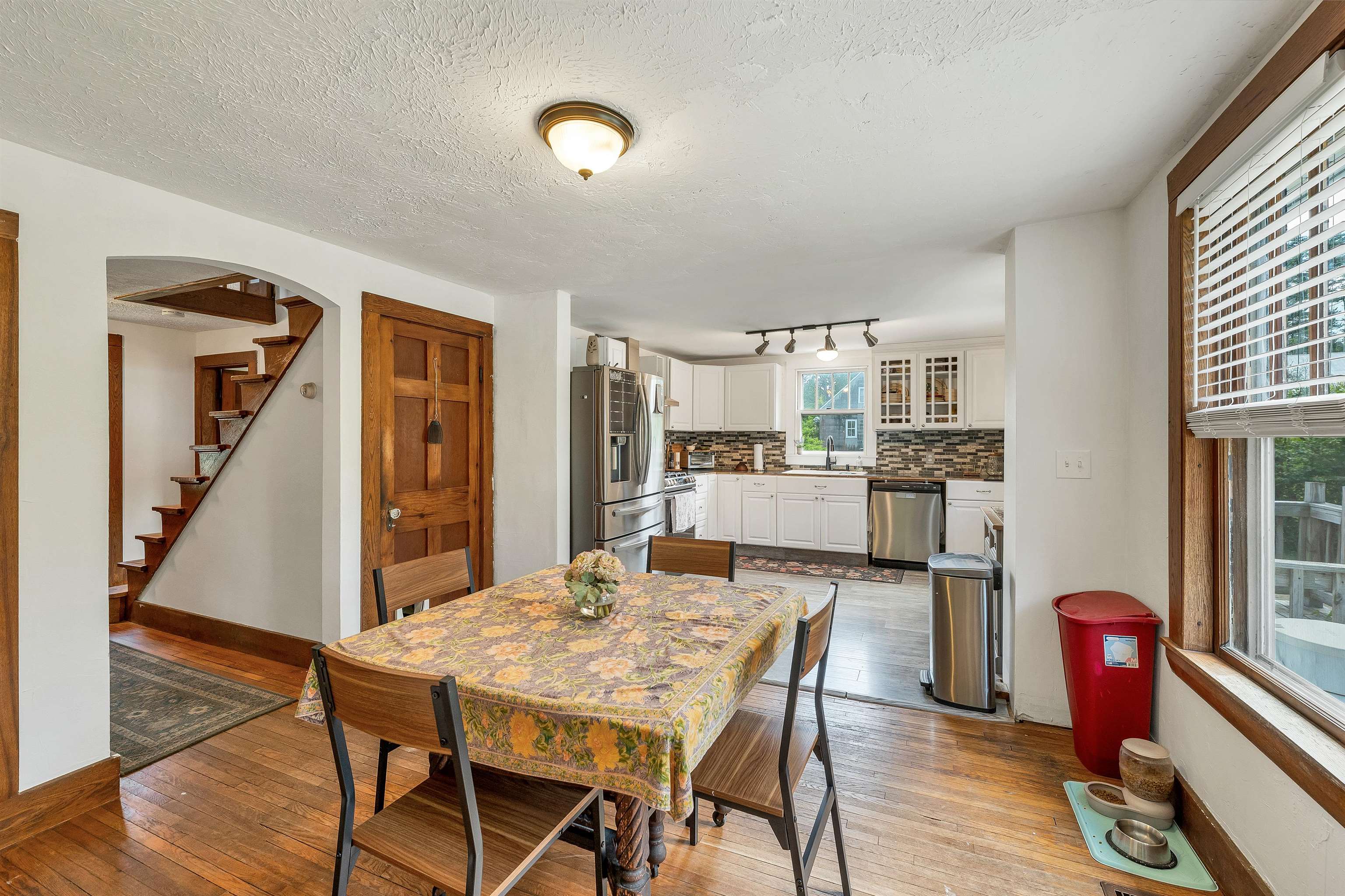Dining area with floral tablecloth beside a white kitchen, stainless appliances, and a wooden staircase.