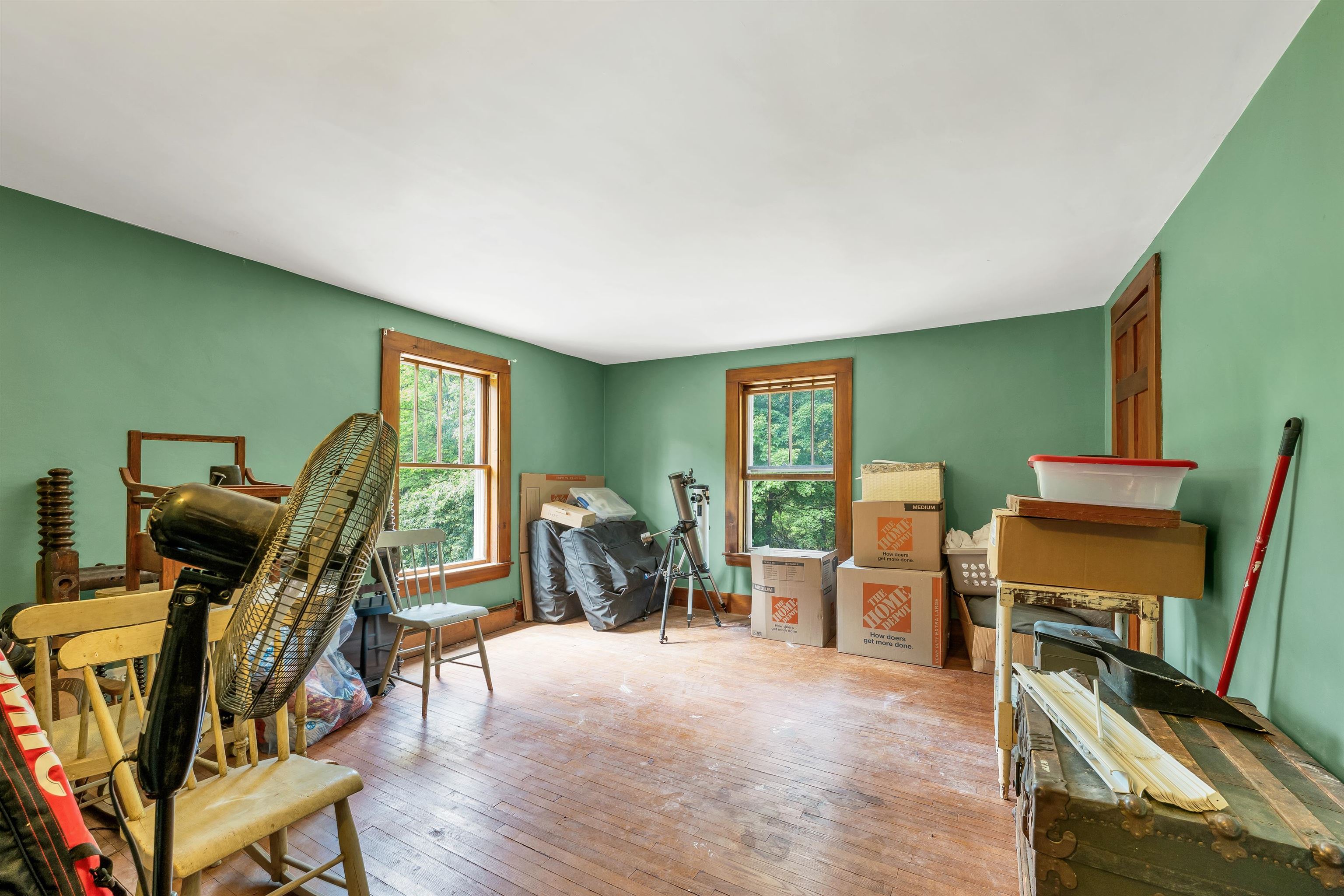 Green-walled room with hardwood floors, two windows, and boxes and furniture stored around the edges.