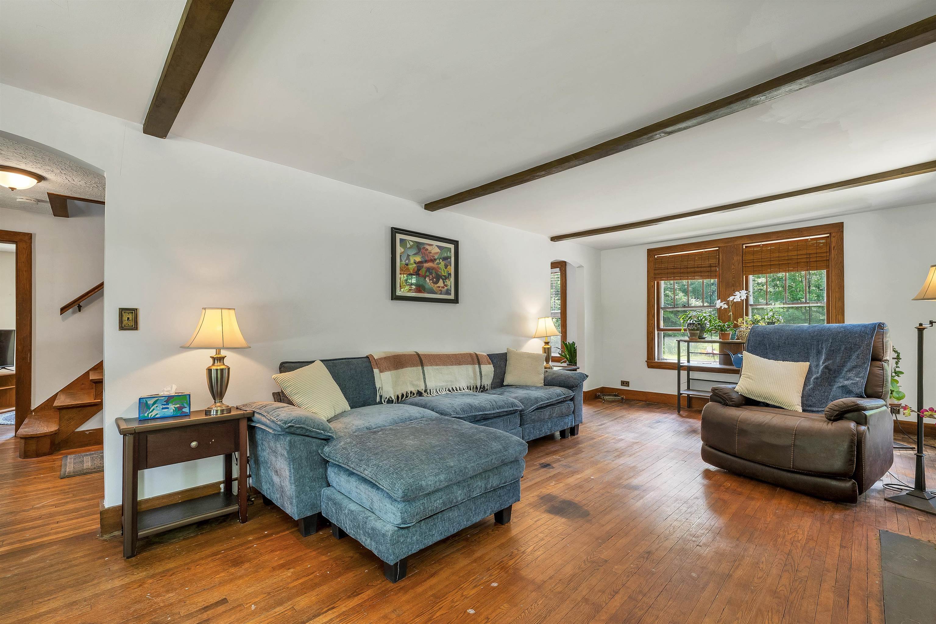 Living room with a blue sectional sofa, hardwood floors, exposed beams, and a large window.