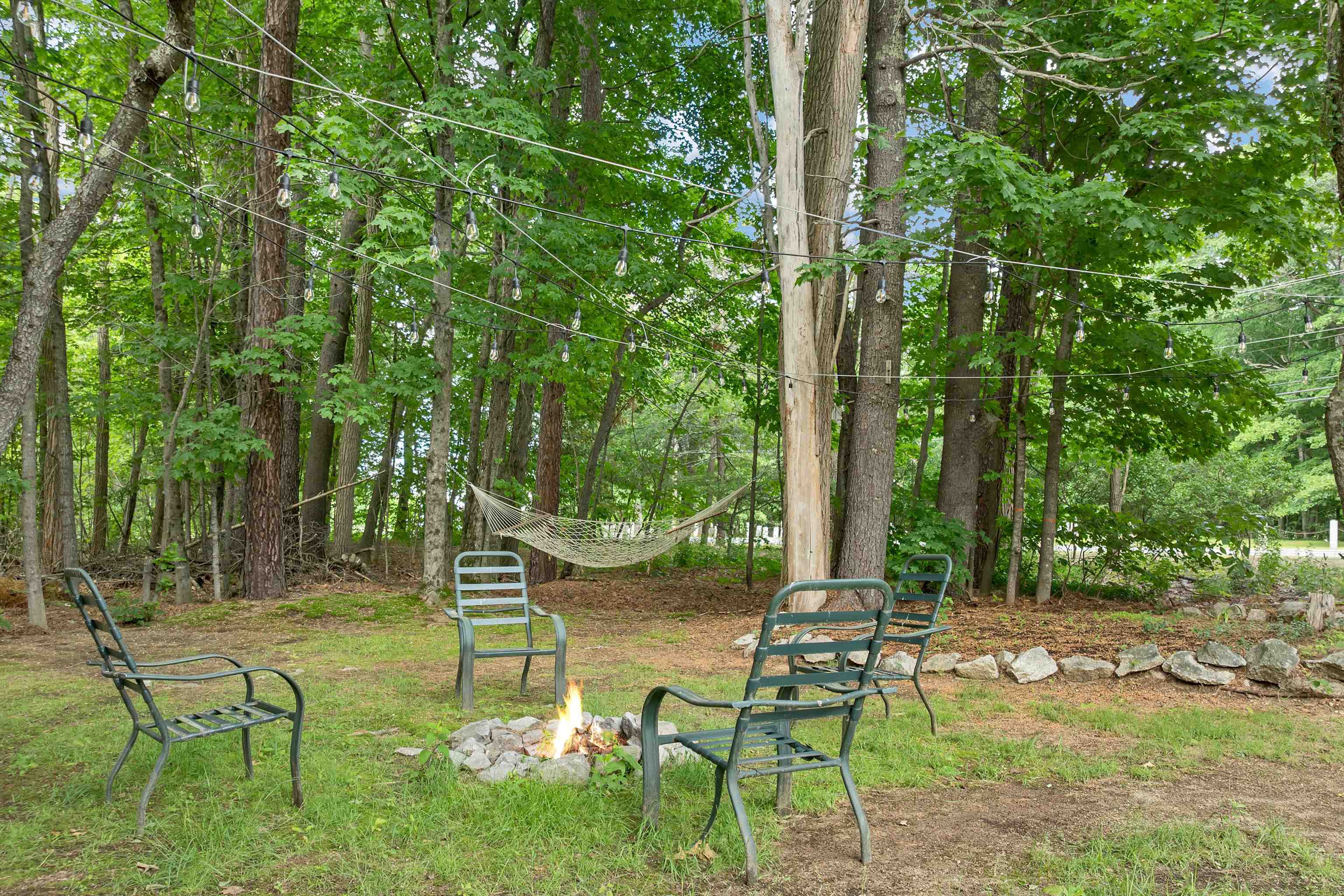 Wooded backyard with a fire pit, metal chairs, hammock, and overhead string lights.