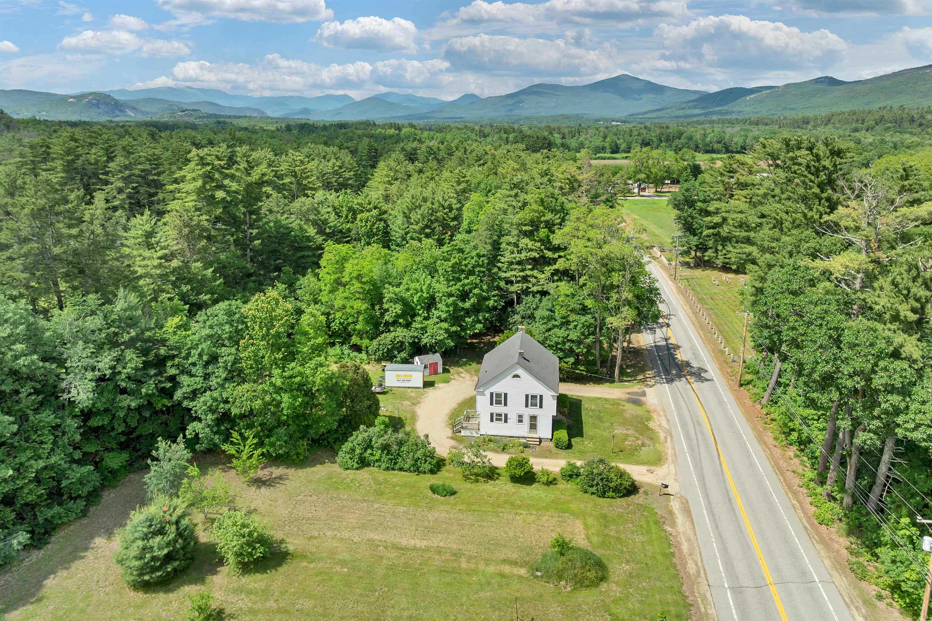 Aerial view of a white house beside a road, with wooded grounds and mountains beyond.