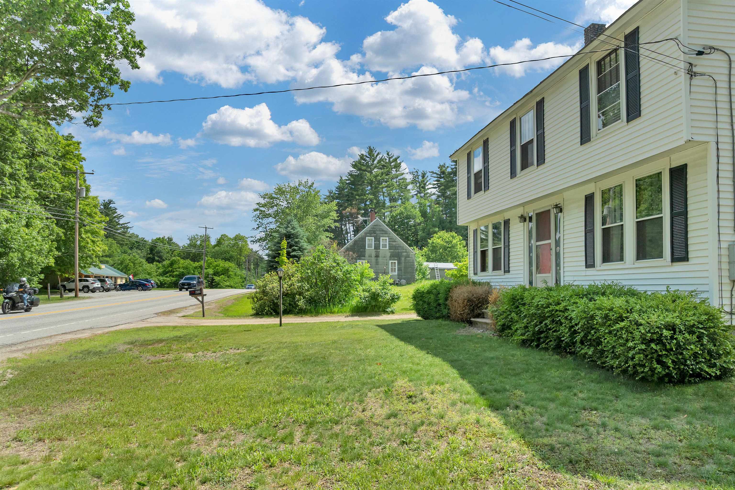 Two-story white-sided home with black shutters, front shrubs, and a lawn beside the road.