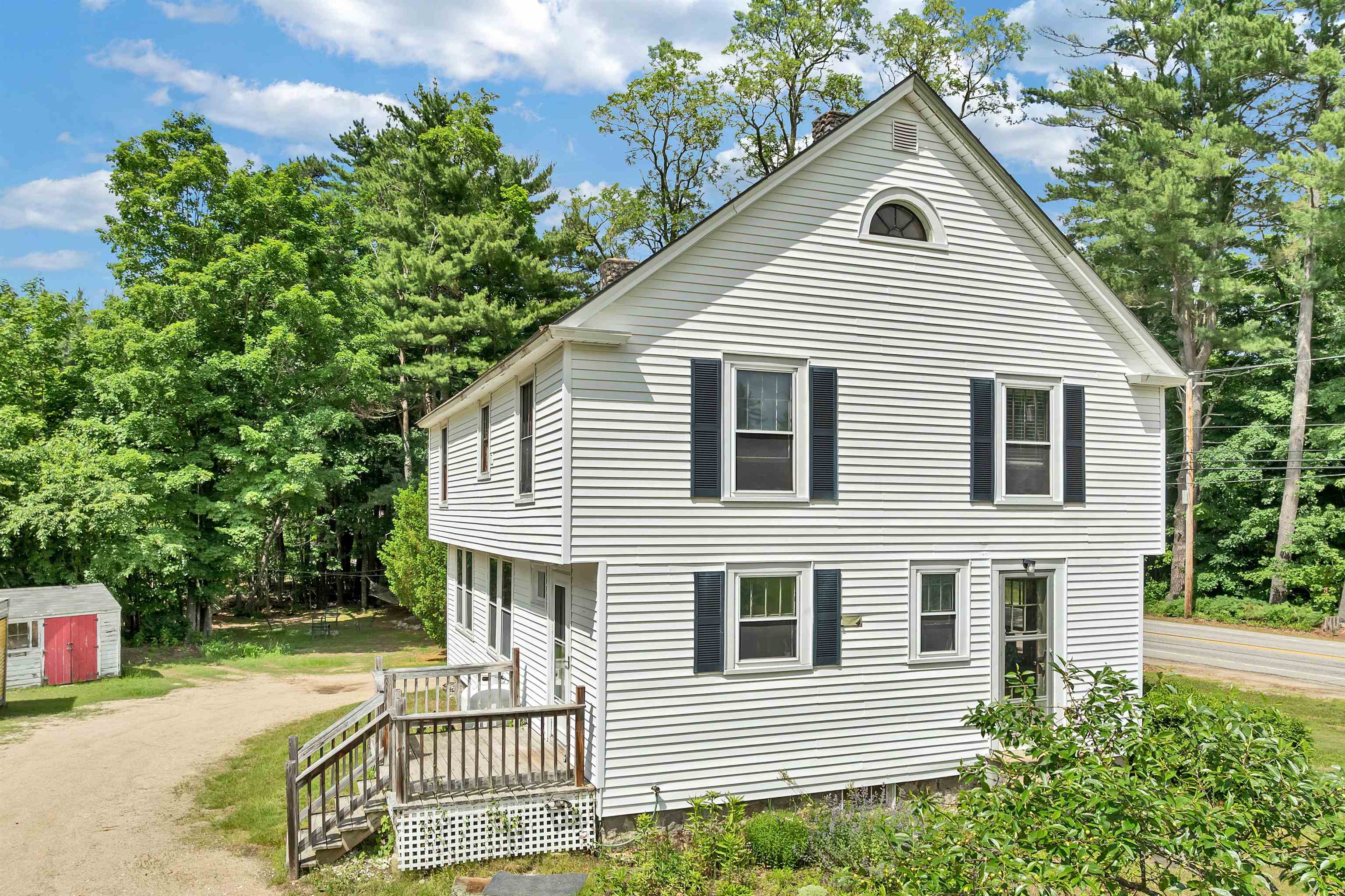 Two-story white-sided house with black shutters and a small wooden porch beside a gravel driveway.