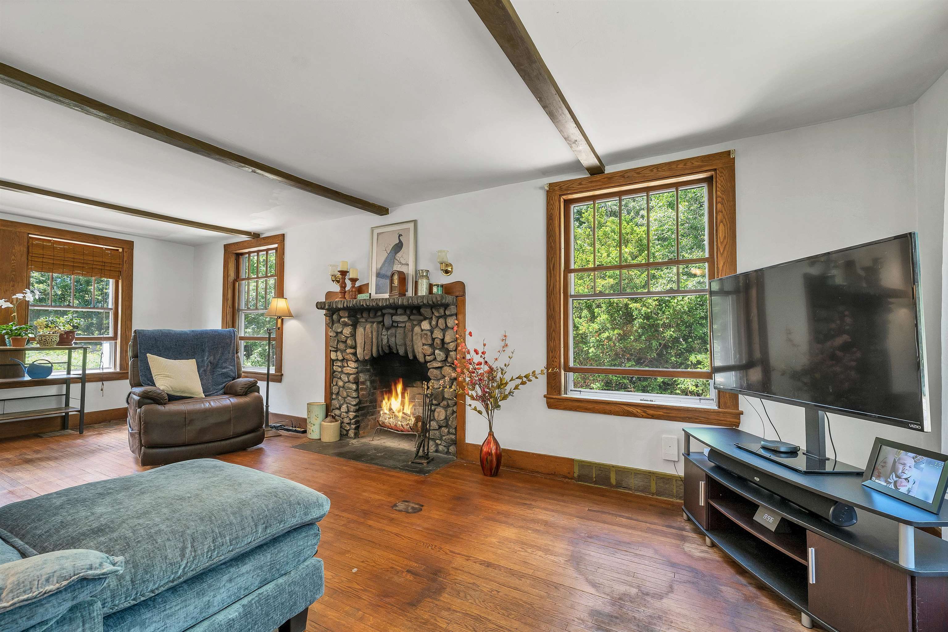 Living room with a stone fireplace, exposed beams, and large wood-trimmed windows.