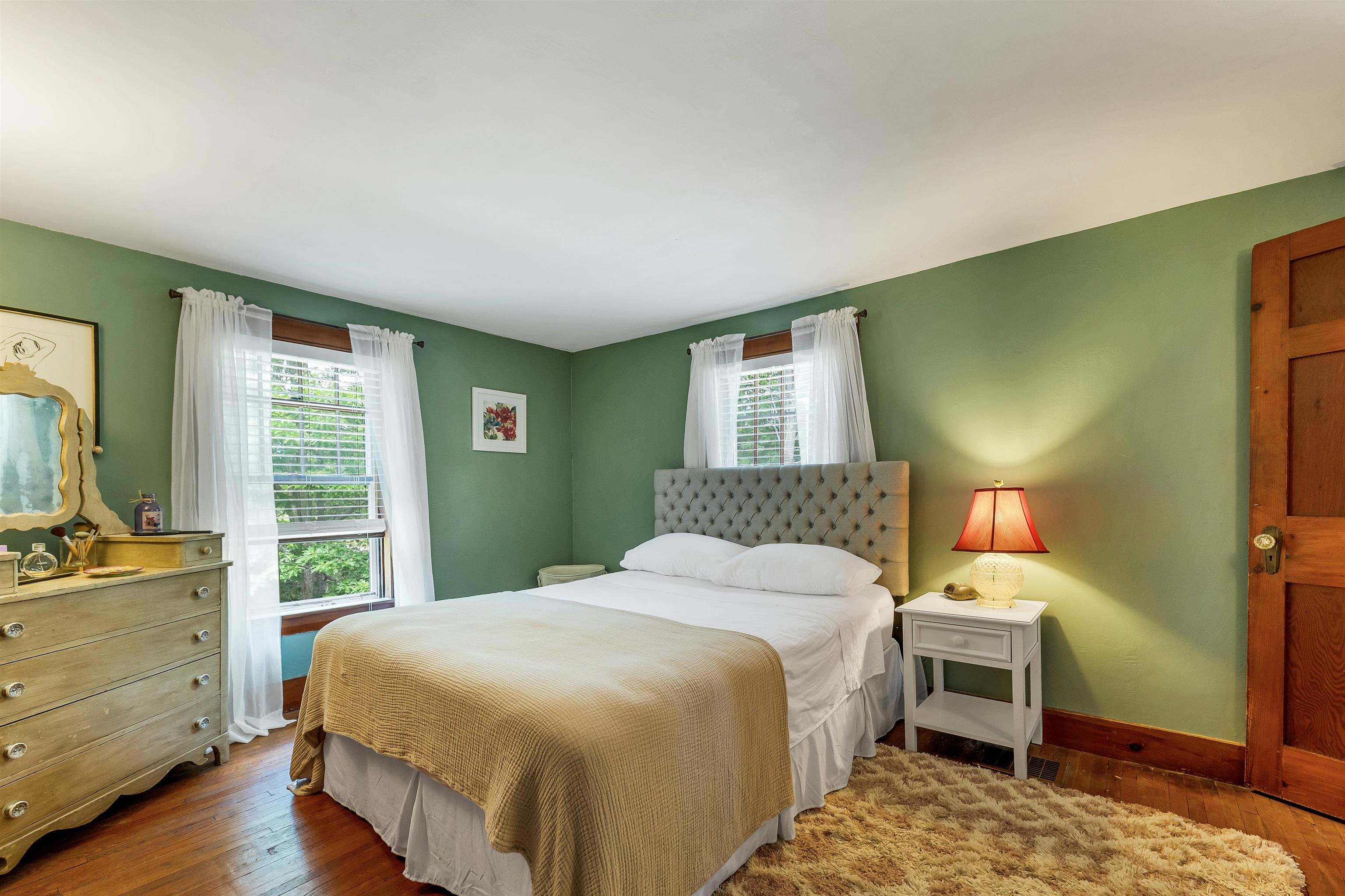 Bedroom with sage-green walls, a white bed, tufted headboard, and two curtained windows.