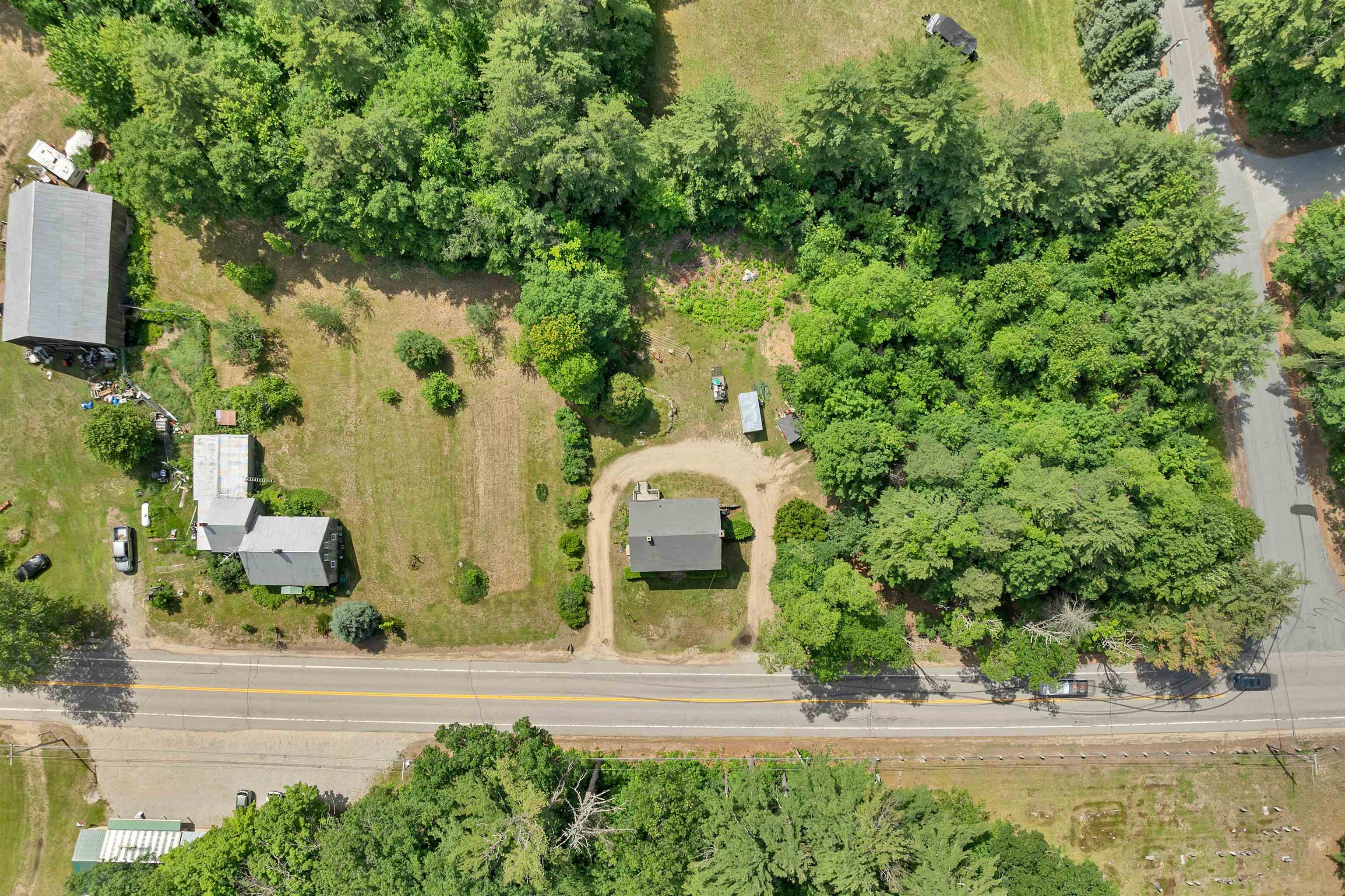 Aerial view of a wooded property with several buildings and a curved gravel driveway beside a road.