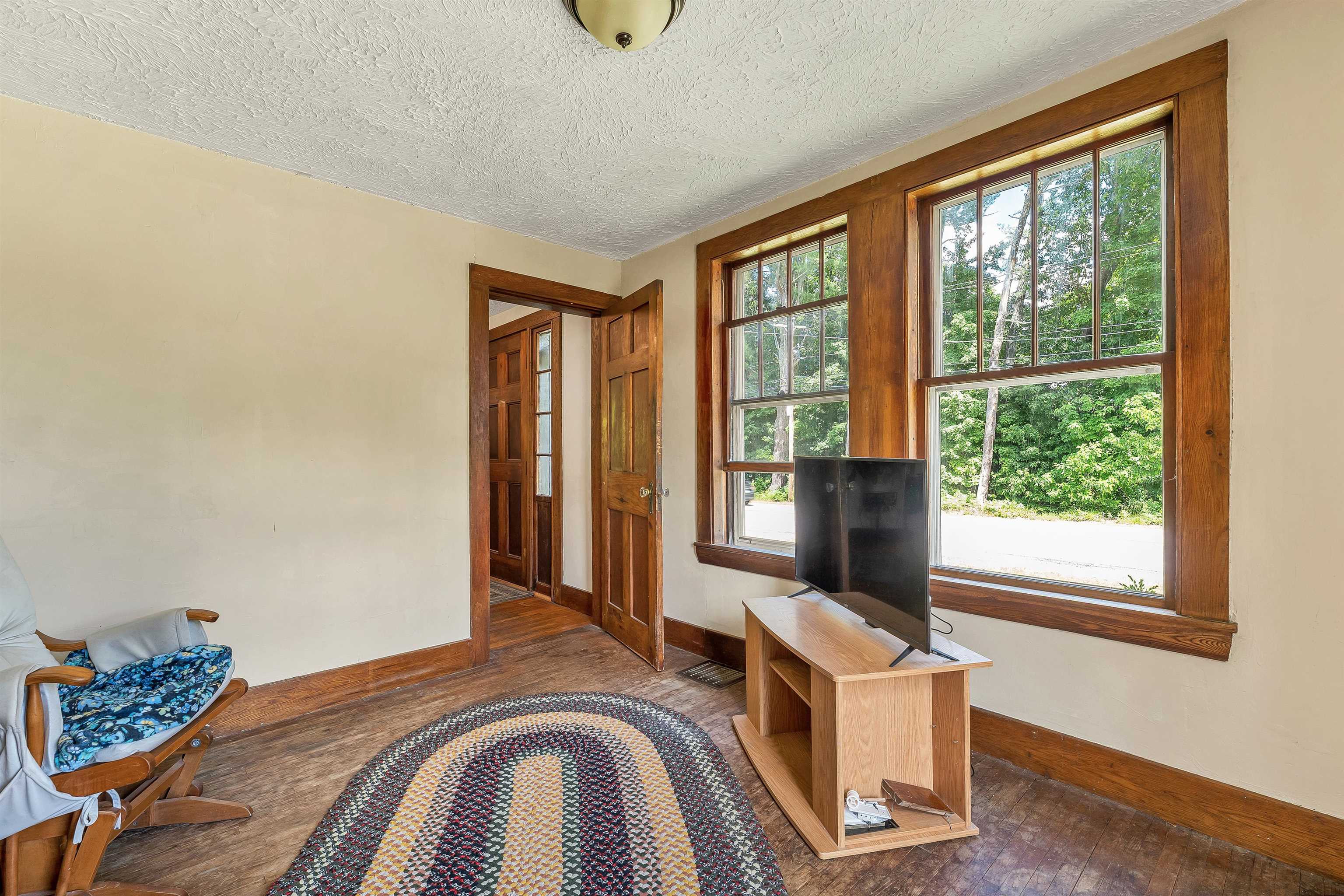 Room with wood-trimmed windows, a television, rocking chair, and braided rug.