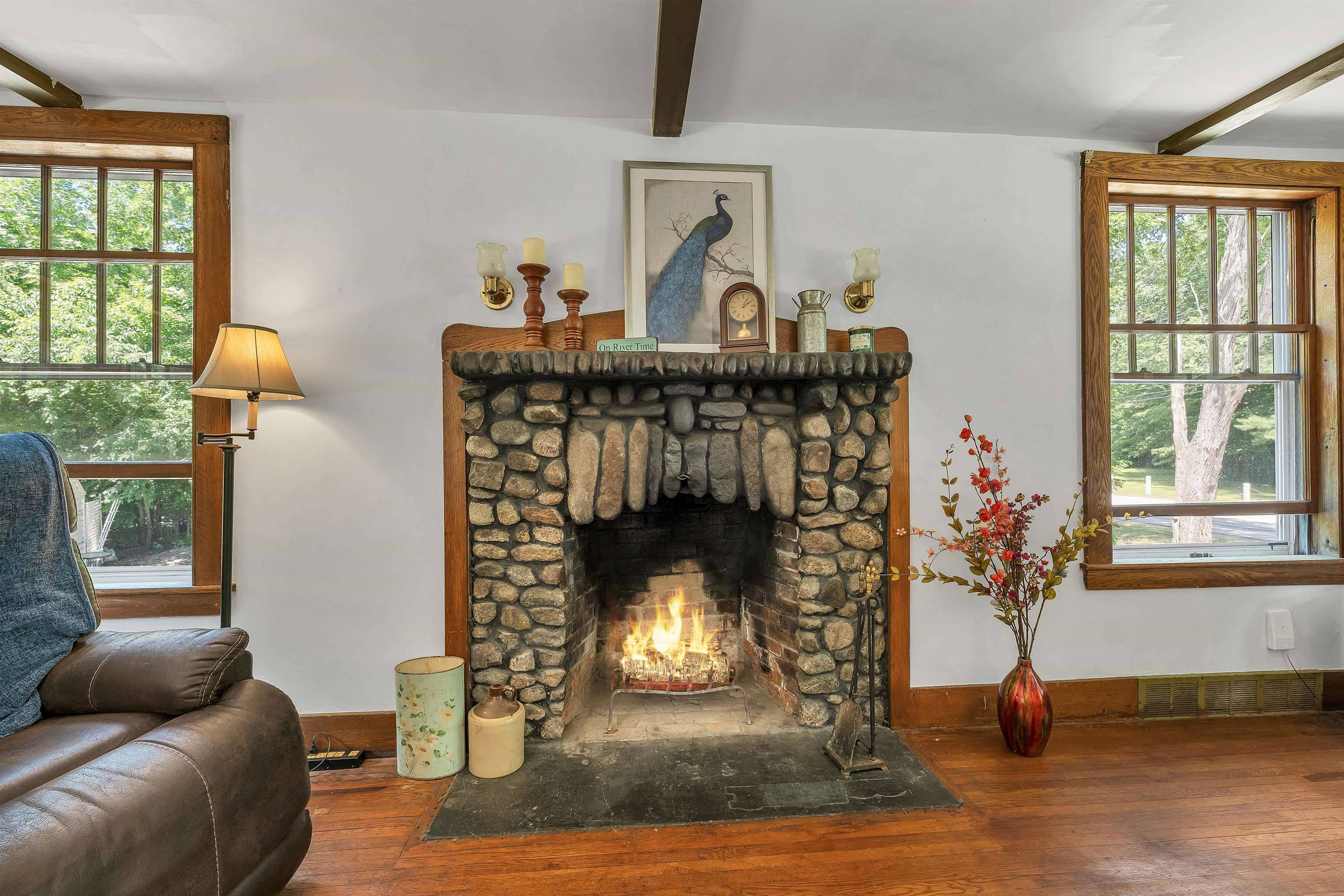 Living room with a large stone fireplace, burning fire, and wood-trimmed windows overlooking greenery.