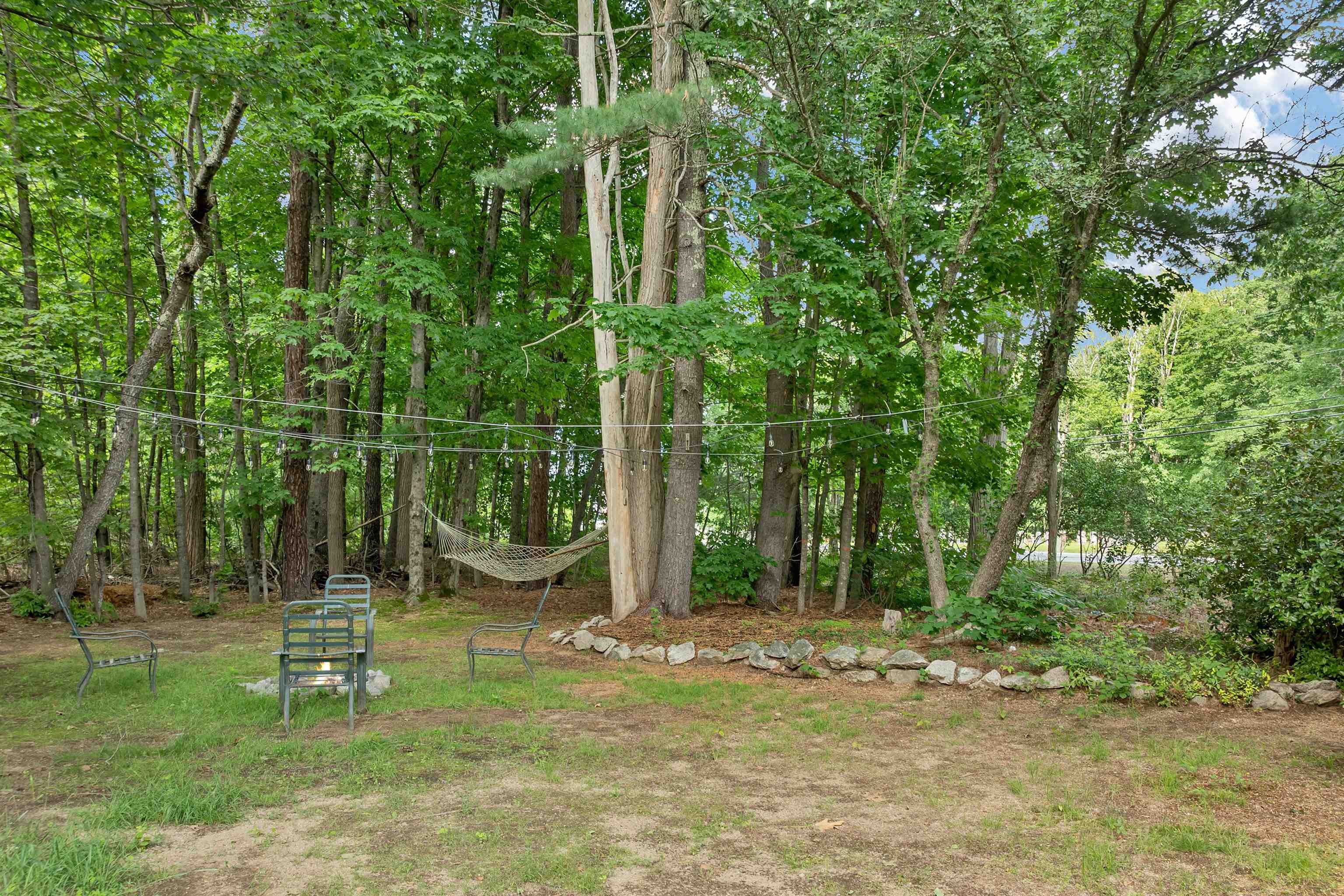 Wooded backyard with a hammock, string lights, outdoor seating, and a small fire pit.