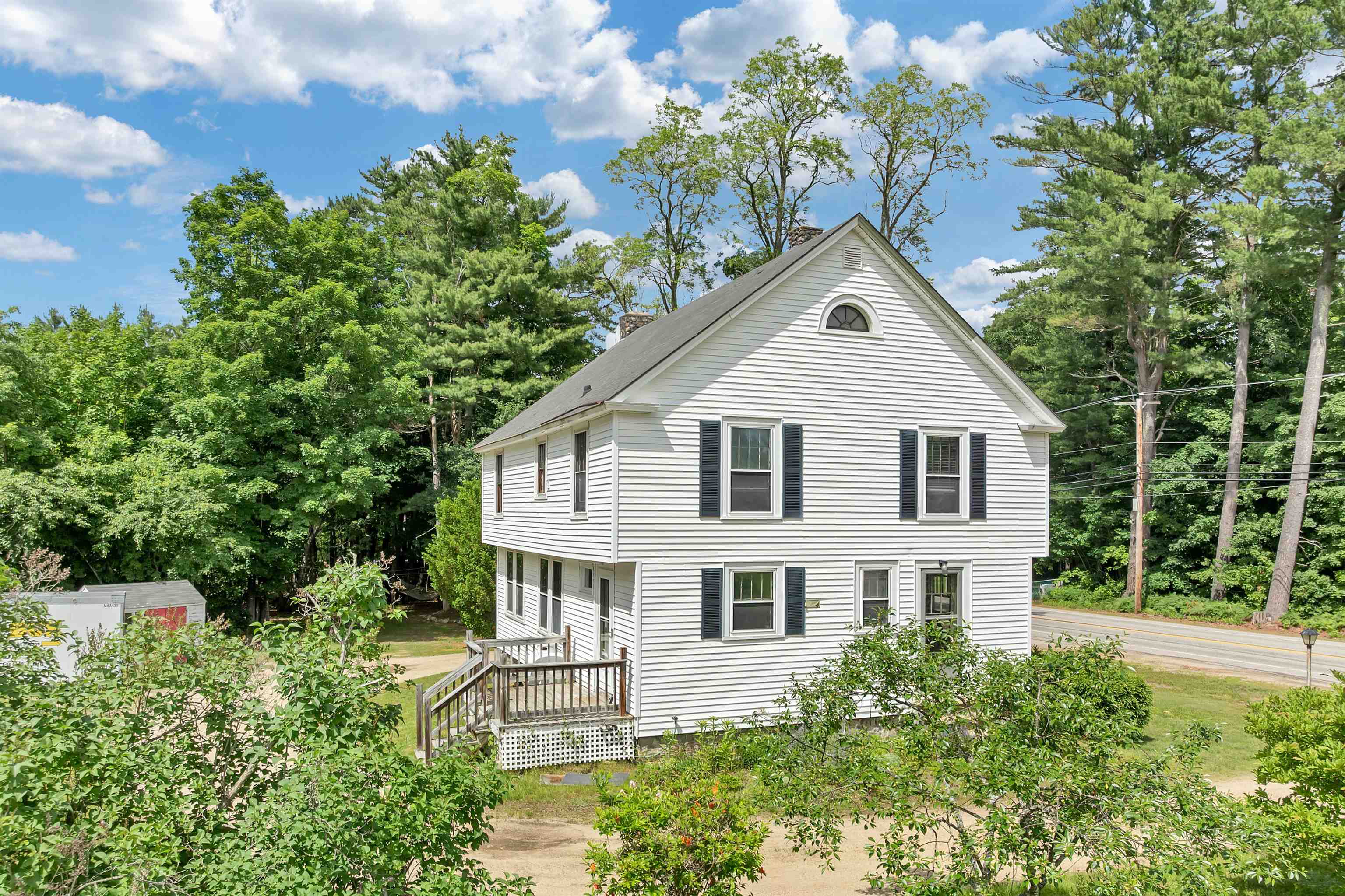 White two-story gabled house with dark shutters and a small wooden porch, surrounded by summer greenery.