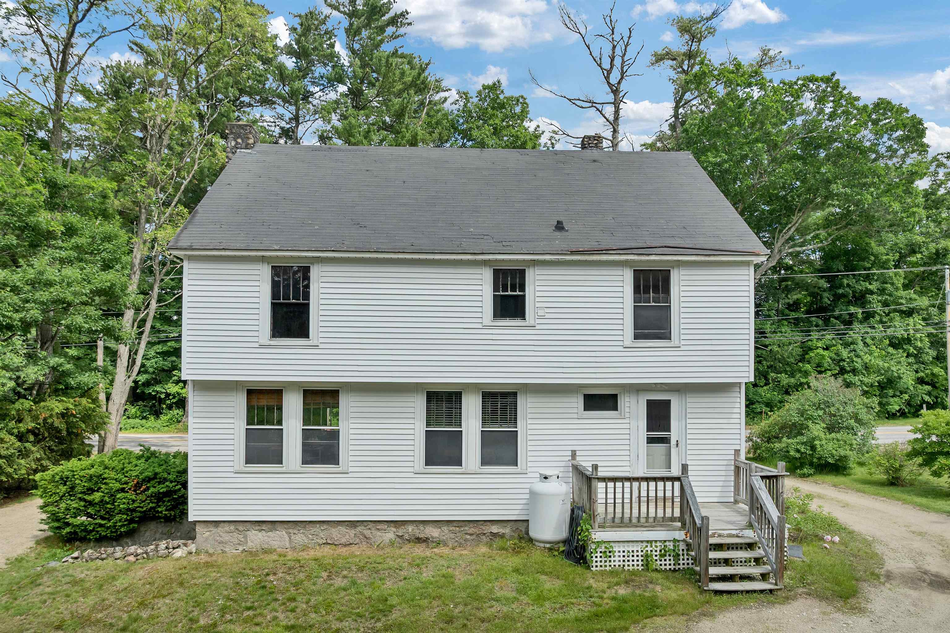 White two-story house with a raised wooden deck and stairs, surrounded by mature trees.