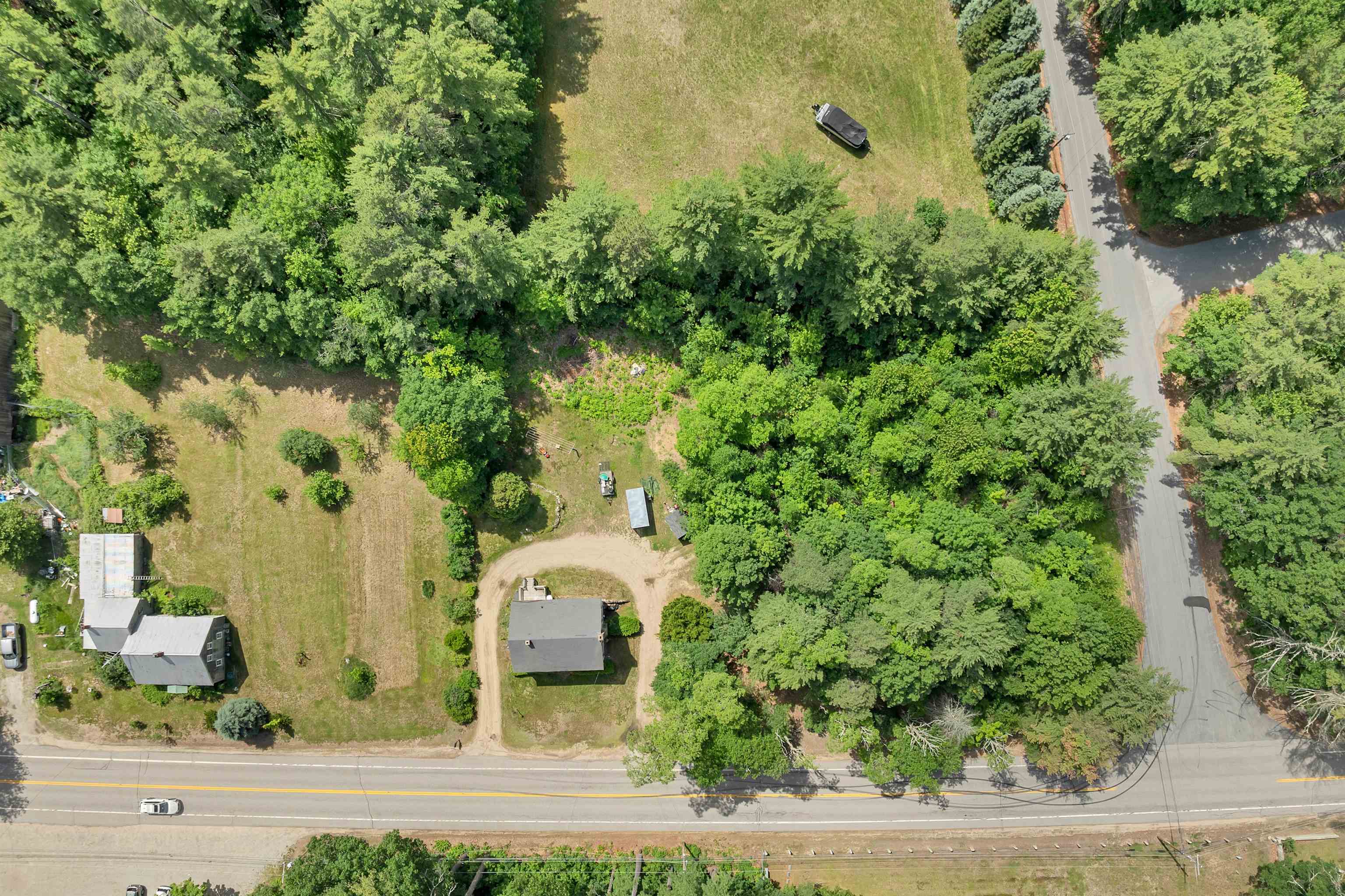 Aerial view of a wooded property with two buildings, open lawn, and a curved driveway beside the road.