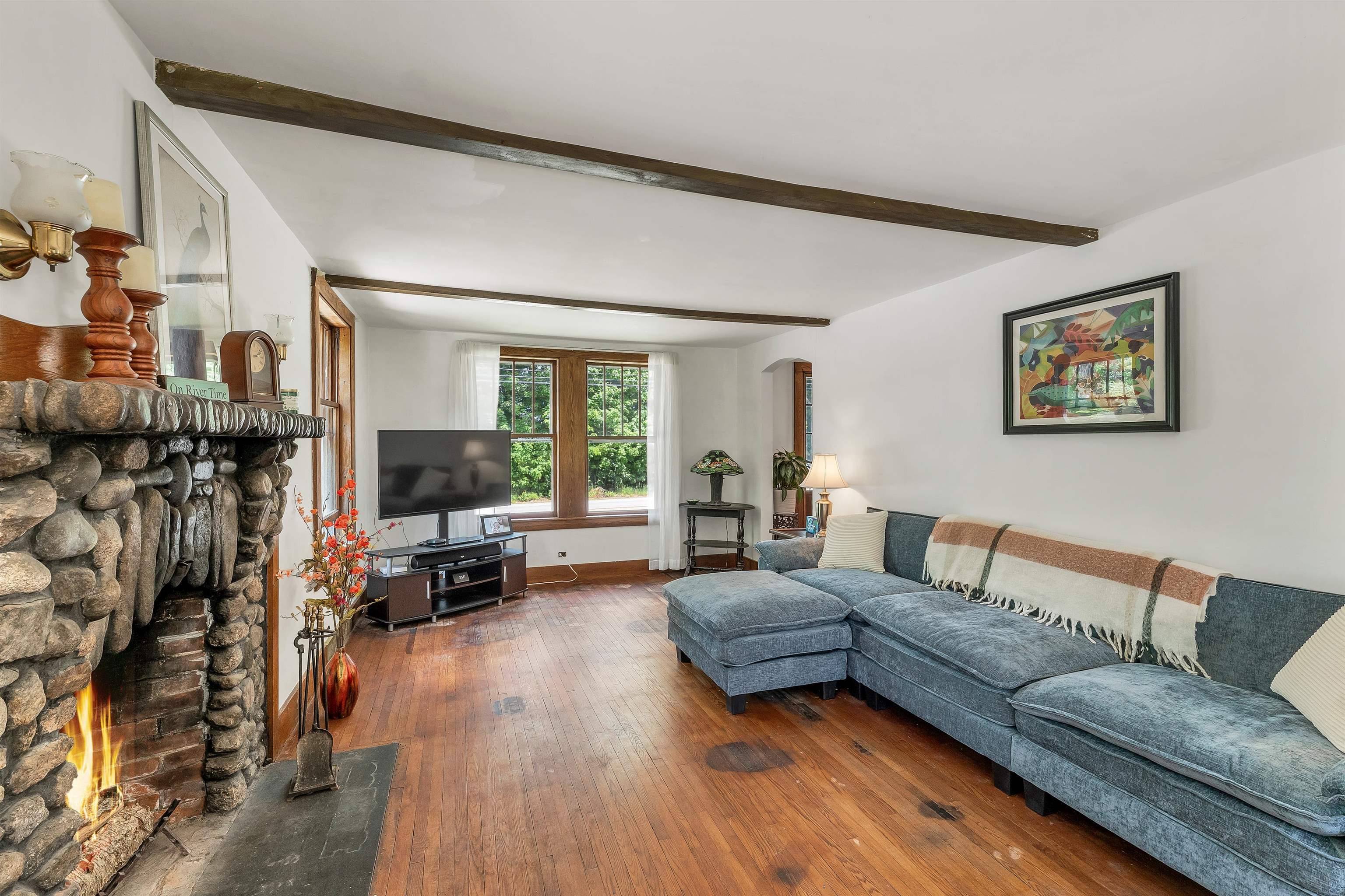Living room with a stone fireplace, exposed ceiling beams, hardwood floors, and a blue sectional sofa.