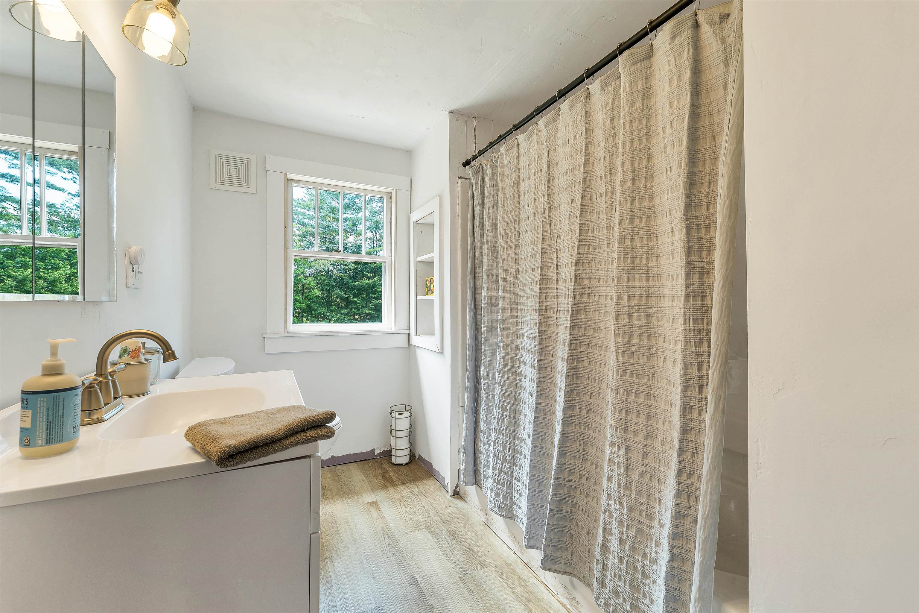 Bathroom with a white vanity, brass-tone faucet, wooded window view, and textured beige shower curtain.