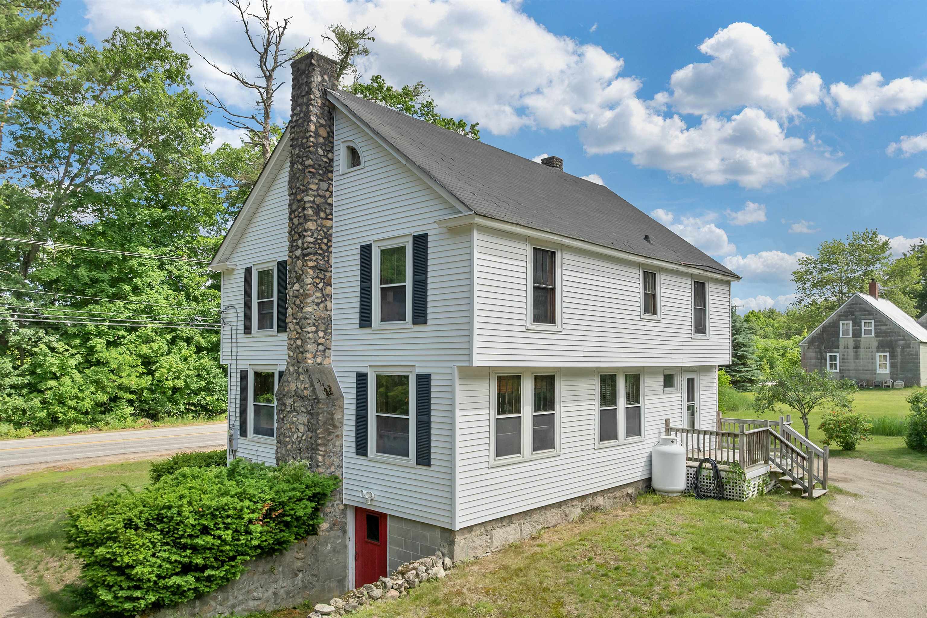 White-sided house with a tall stone chimney, black shutters, and red basement door beside a road.