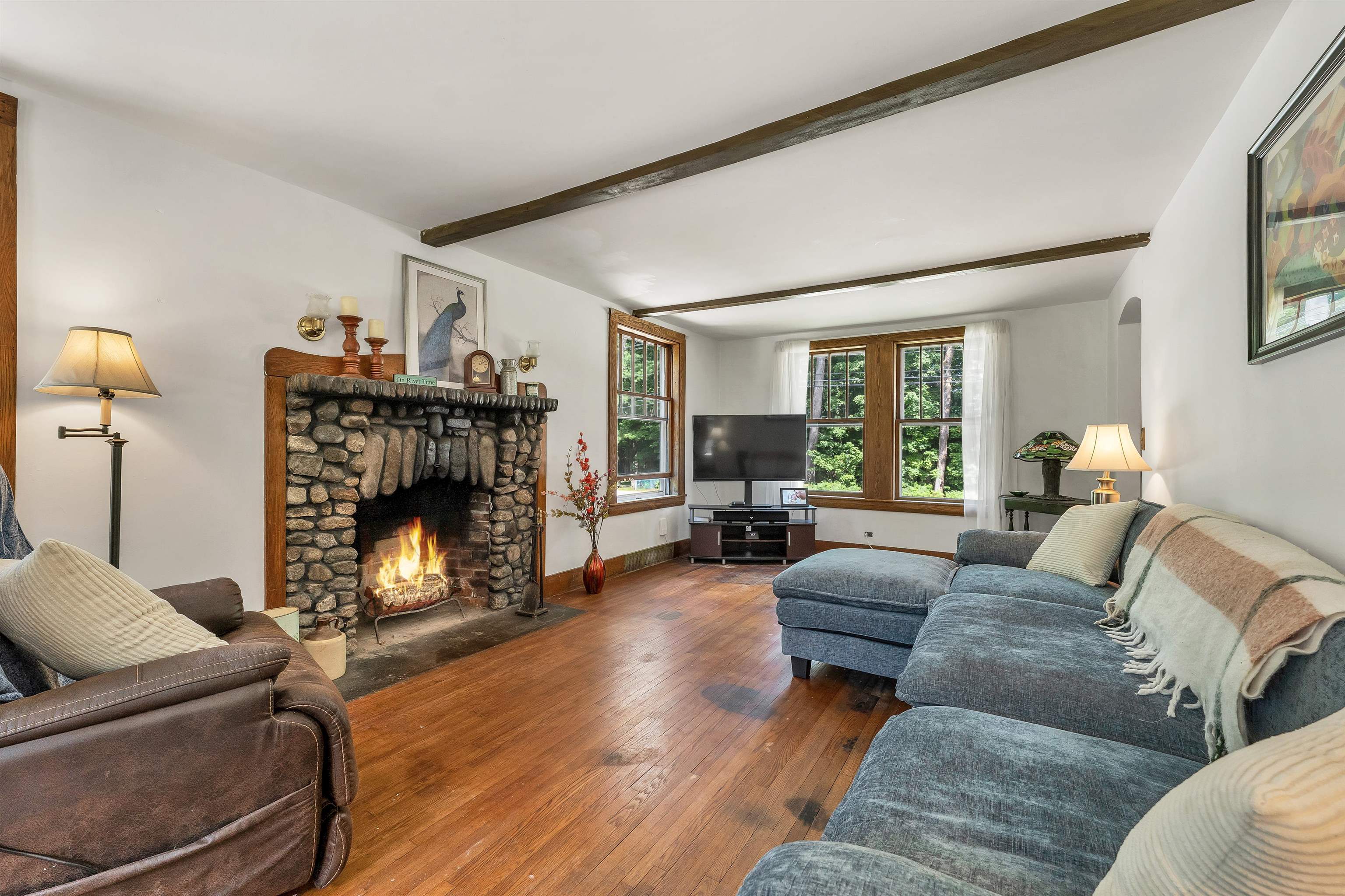 Living room with a stone fireplace, blue sectional sofa, exposed beams, and windows overlooking trees.