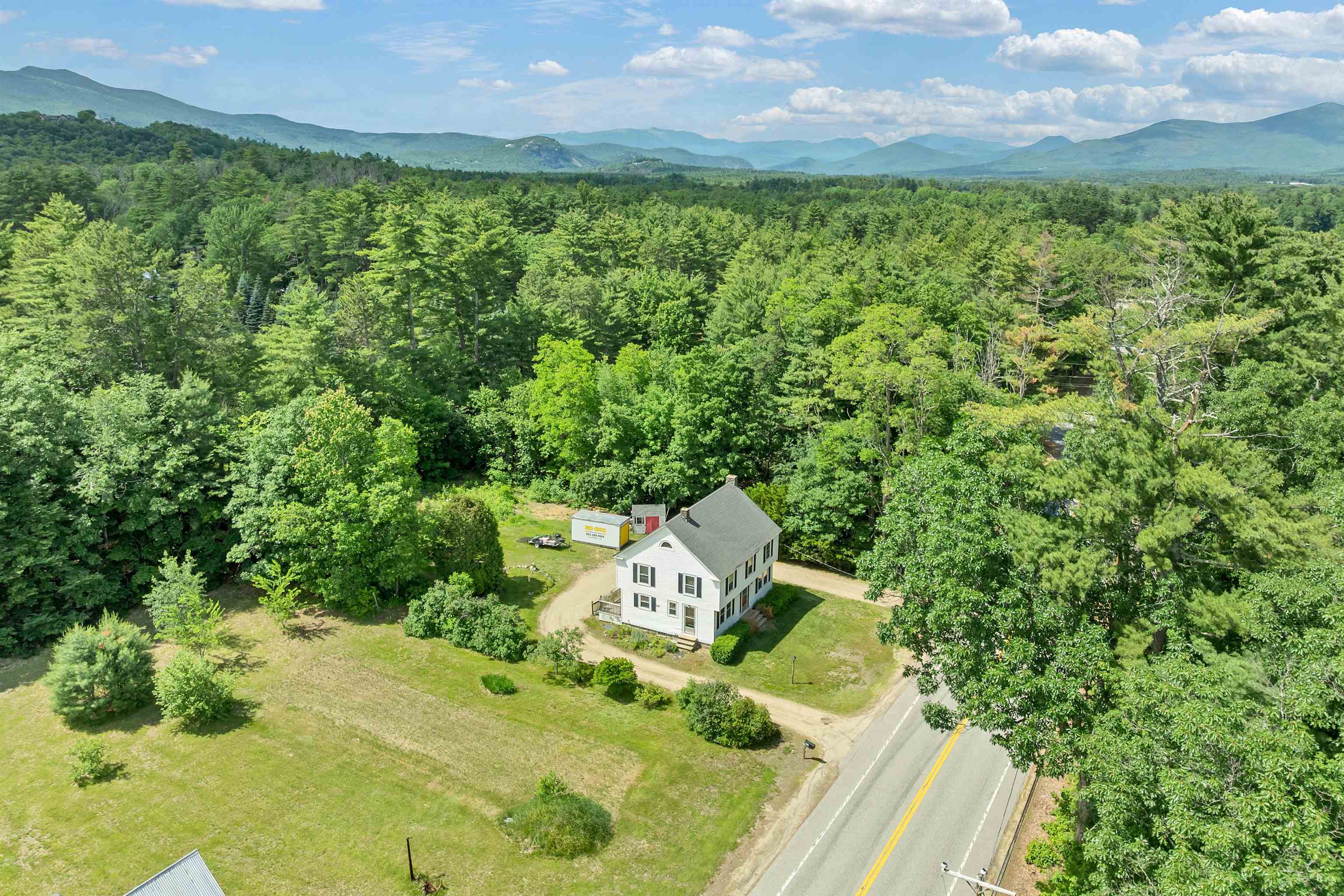 Aerial view of a white two-story house beside a road, with wooded grounds and mountains beyond.