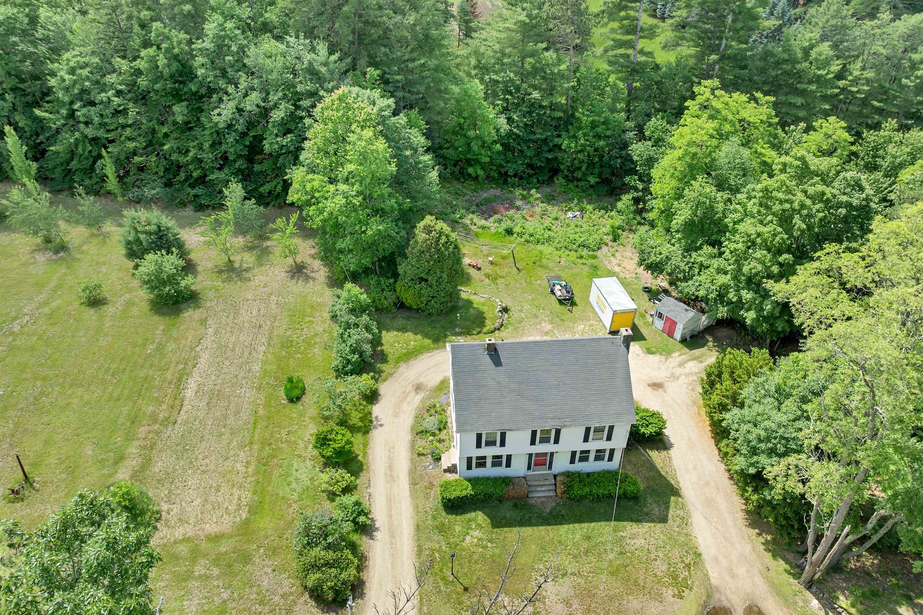 Aerial view of a white two-story house with a gravel driveway, grassy yard, shed, and surrounding trees.