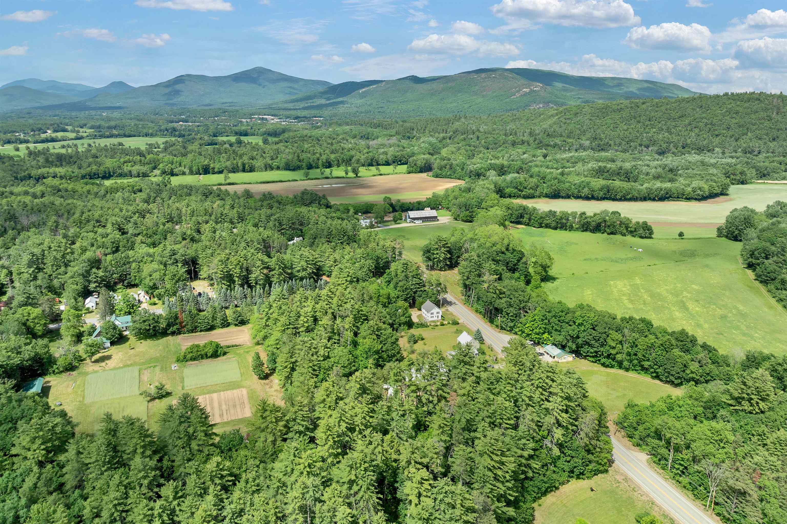 Aerial view of wooded countryside with open fields, a road, scattered homes, and distant mountains.